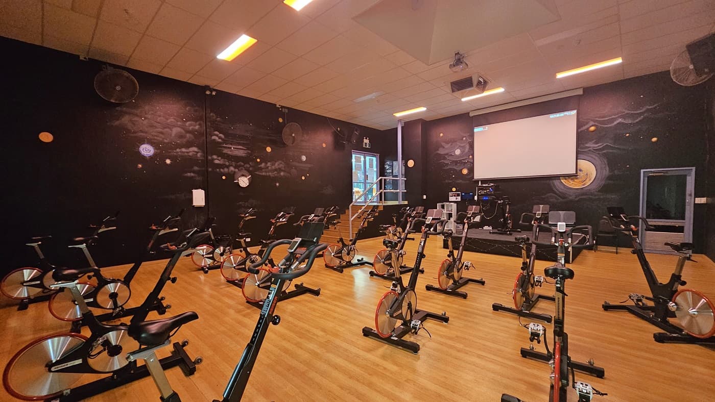 Spin bikes are arranged on the timber floor. A rubbish bin is placed near the right wall. The surrounding wall is black with space imagery. Wall fans are affixed to the upper walls. The ceiling is white with fluorescent lights, featuring a projector and aircon vents. To the right is an elevated platform with a spin bike. At the back is a projector screen, and to its left is a staircase.