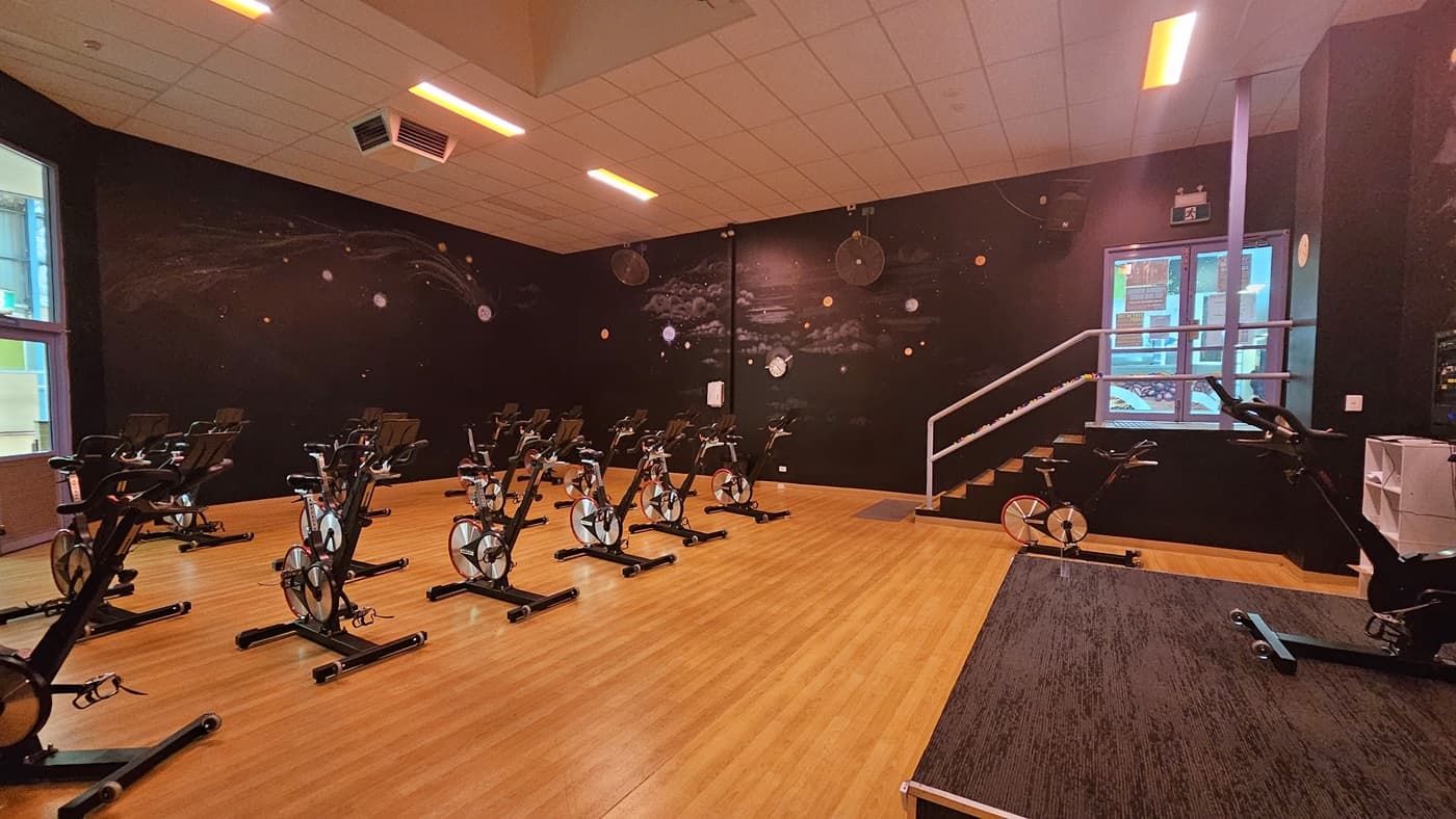 Spin bikes are arranged on the timber floor. The surrounding wall is black with space imagery. Wall fans are affixed to the upper walls. The ceiling is white with fluorescent lights, featuring a projector and aircon vents. To the right is an elevated platform with a spin bike. Adjacent is a white display rack screen, and to its left is a staircase.