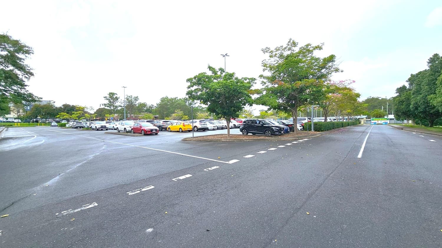 A large car park with numerous vehicles, including a red car and a yellow car, is visible in the centre. Trees are scattered throughout, providing some greenery. The road in the foreground has dashed white lines, leading towards the car park. On the right, a road extends into the distance, bordered by more trees. The background features more trees and a few buildings partially visible. The sky is overcast.