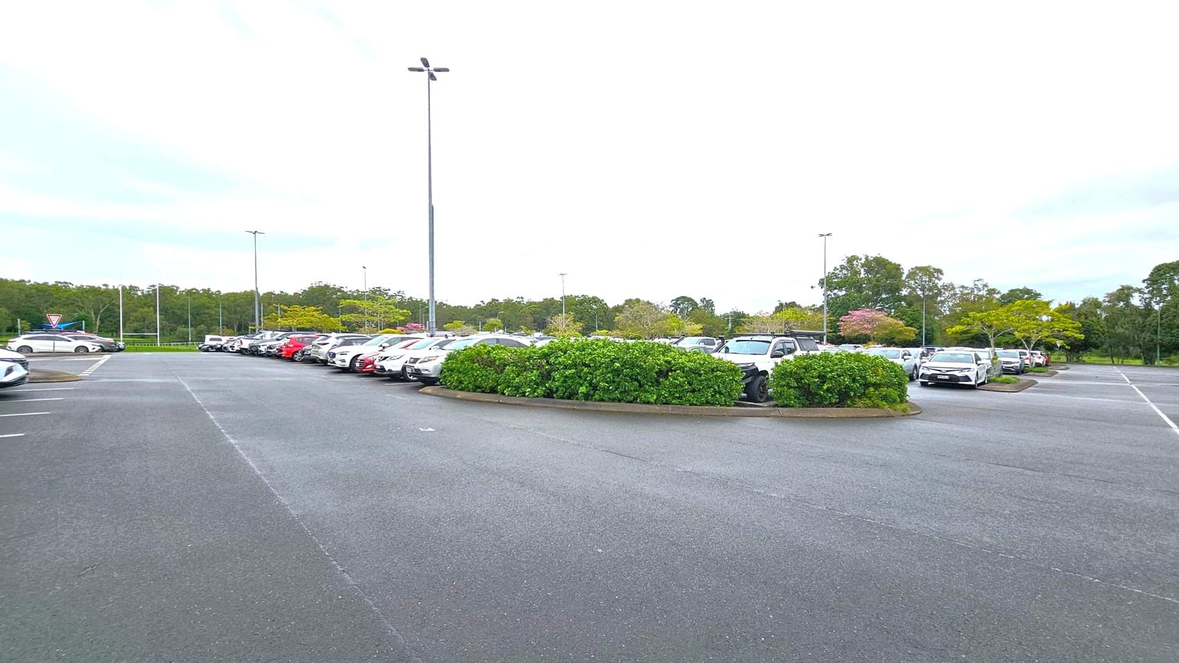 A car park with multiple vehicles parked in rows. In the centre, there is a landscaped area with green bushes. The ground is paved with asphalt, and tall streetlights are visible. In the background, there are trees and greenery, providing a natural border to the parking area.