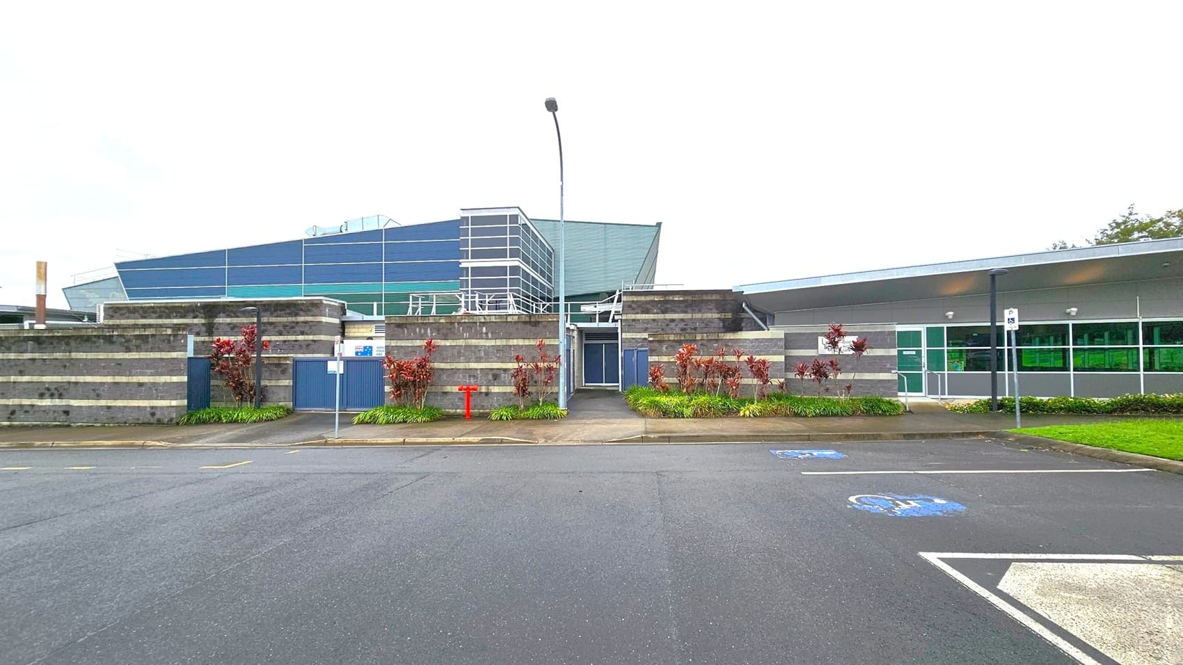 A modern building with a mix of grey and green panels. The left side features a structure with horizontal stripes and a central entrance, flanked by small plants. The right side has large windows and a flat roof. In the foreground, there is a paved area with marked parking spaces, including a disabled parking spot. A tall streetlamp stands in front of the entrance. The surrounding area is landscaped with grass and shrubs.