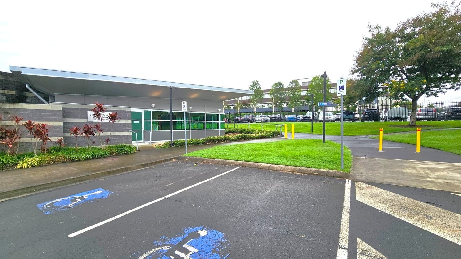 A modern building with a flat roof is on the left, featuring grey and green panels and a sign. In front, there are landscaped plants and a pathway. The foreground shows a car park with a marked disabled parking space. On the right, a grassy area with yellow bollards and a paved path leads to a fence. Beyond the fence, there are parked cars and trees. The sky is overcast.