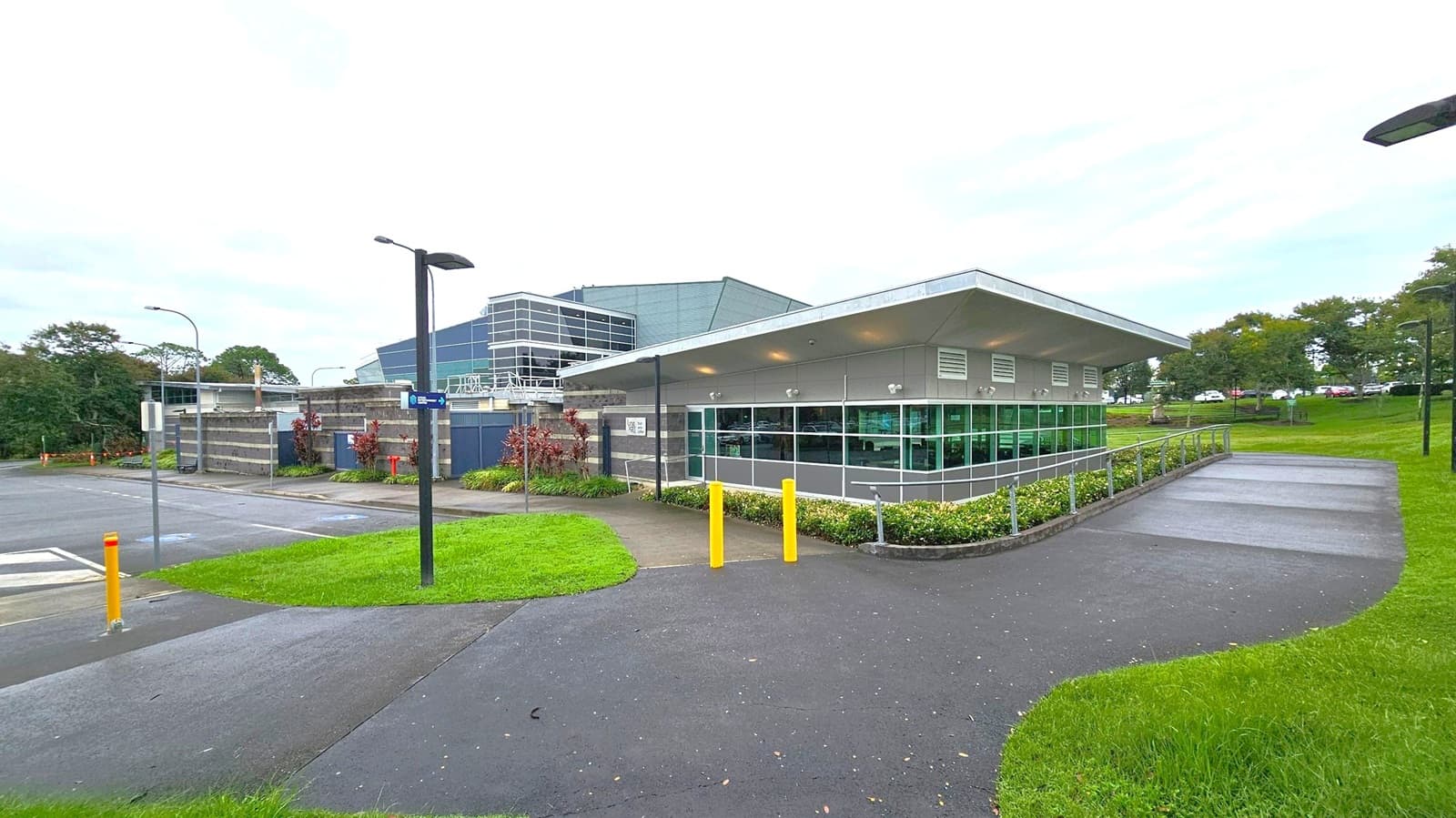 A modern building with large glass windows and a sloped roof is situated on the right. It is surrounded by a well-maintained lawn and a paved path with yellow bollards. On the left, there is a parking area with a few street lamps and signs. In the background, trees and more greenery are visible, along with a larger building with a glass facade. The sky is overcast, and the ground is wet, suggesting recent rain.