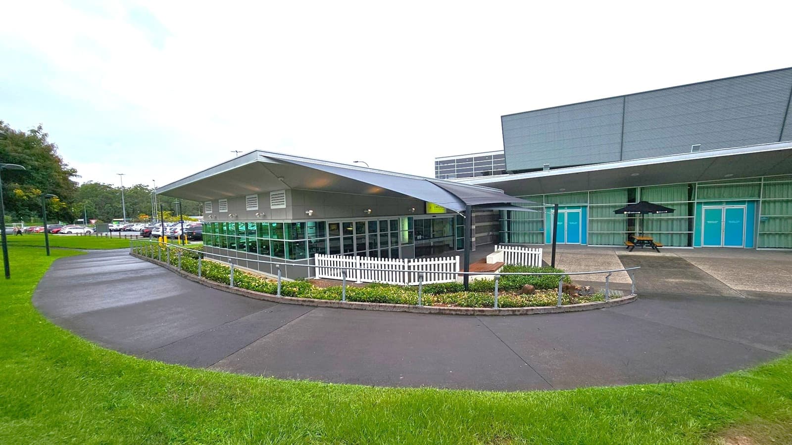 A modern building with a sloped roof and large glass windows on the left. A white picket fence surrounds a small garden area. The right side features a larger structure with blue doors and a covered area with a picnic table and umbrella. The foreground has a curved pathway bordered by green grass, leading to a car park visible in the background.
