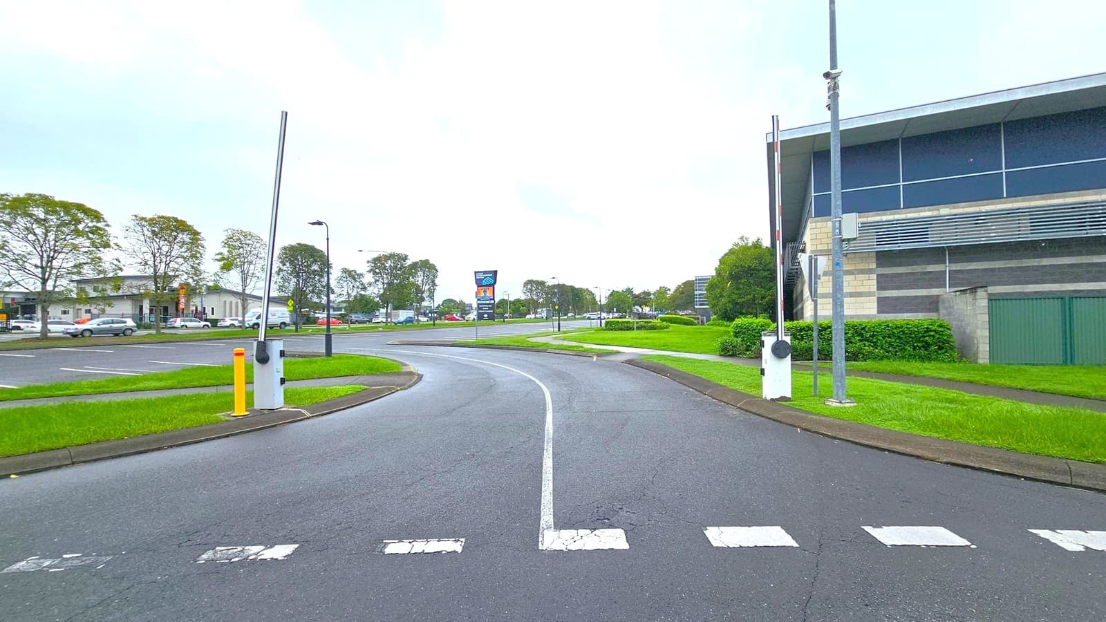 A road with a central lane curves into the distance, flanked by grassy verges. On the right, a modern building with large windows and a green fence is visible. A tall pole with a barrier arm stands nearby. On the left, a parking area with several cars and a row of trees can be seen. In the background, a large sign and more trees line the road. The sky is overcast, creating a muted atmosphere.