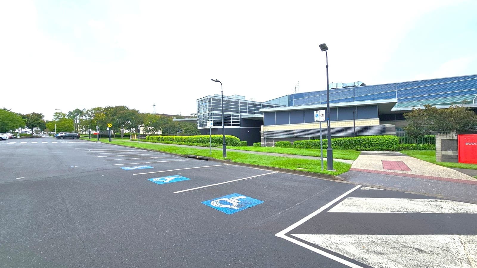A parking area with marked spaces, including disabled parking spots with blue symbols on the asphalt. On the right, a modern building with large glass windows and a flat roof is visible, surrounded by neatly trimmed hedges. A pathway with a red and beige pattern leads to the building. Street lamps line the path, and a red sign is partially visible on the right. Trees and parked cars are on the left, with a road running alongside.