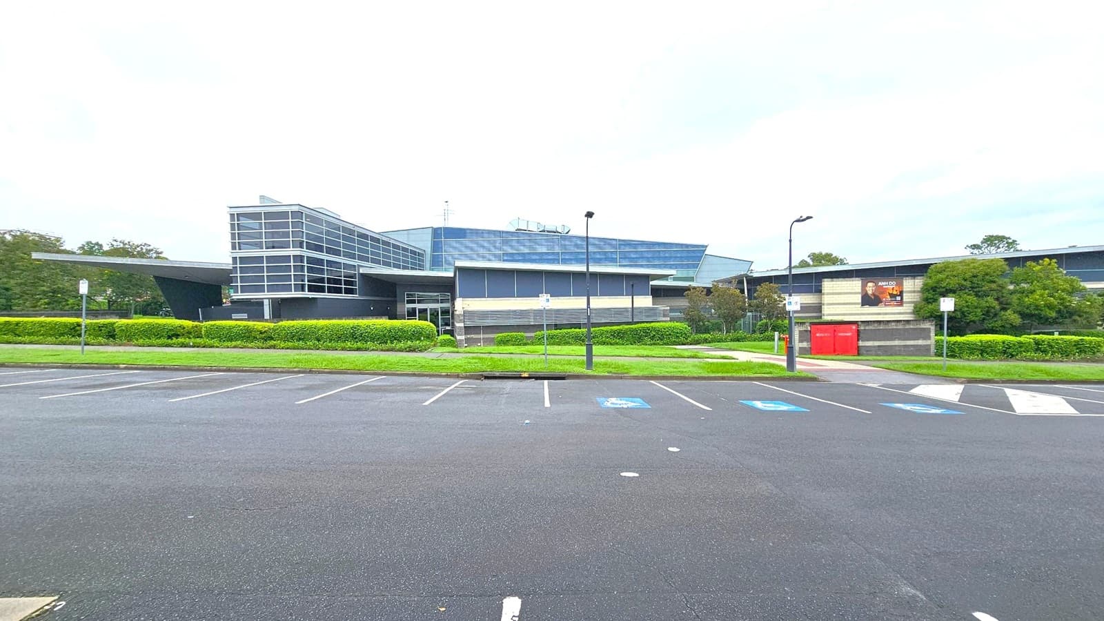 A modern building with a glass facade and angular design is in the centre, surrounded by well-maintained hedges and greenery. To the right, a billboard is mounted on a wall next to red utility boxes. The foreground features an empty car park with marked spaces, including accessible parking spots. Street lamps line the pathway leading to the building.