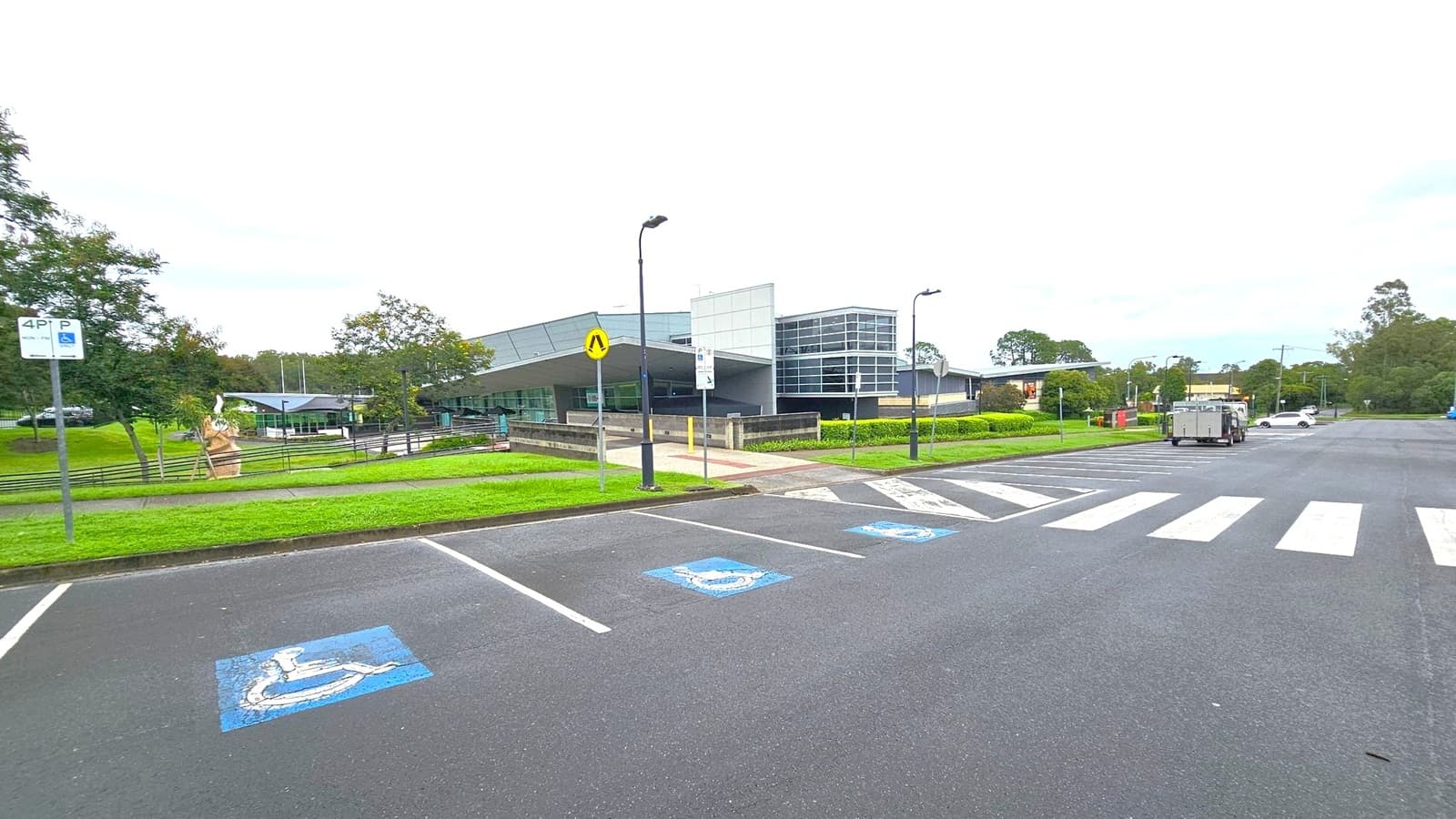 A modern building with large glass windows is in the centre, surrounded by green lawns and trees. On the left, a sculpture is visible near a pathway. The foreground features a car park with marked disabled parking spaces and a zebra crossing. A few street lamps are scattered around, and a white car is parked on the right side. The area is bordered by more greenery and trees in the background.