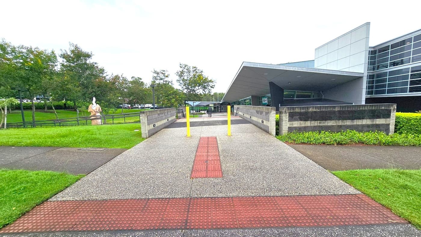 A pathway with a textured red strip runs through the centre, flanked by two yellow bollards. On the right, a modern building with large glass windows and a slanted roof is visible. To the left, a grassy area with trees and a sculpture can be seen. The pathway is bordered by low brick walls and greenery. The sky is overcast.