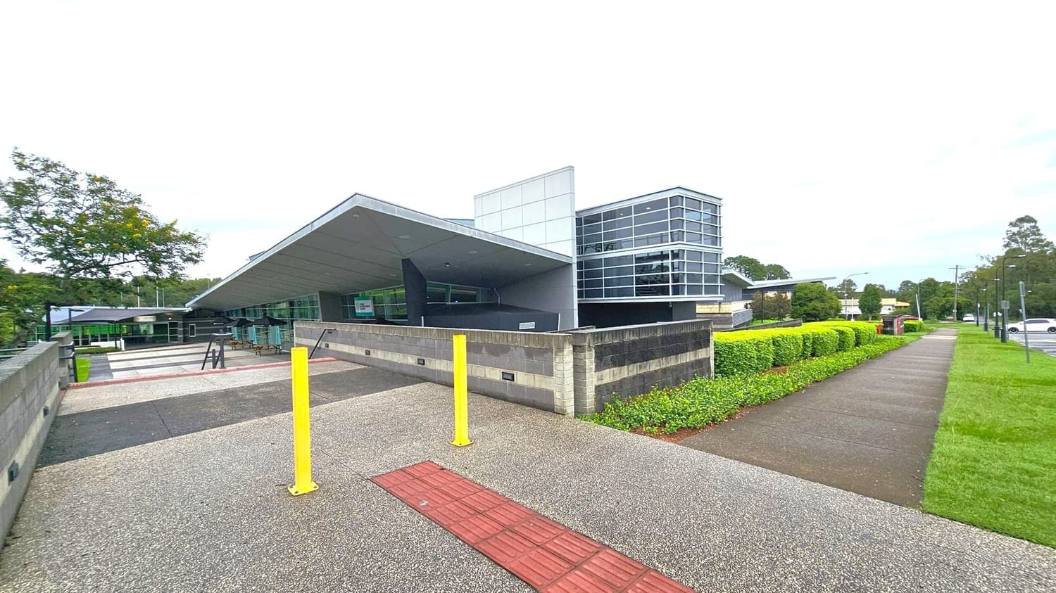 A modern building with a slanted roof and large glass windows is in the centre. The foreground features a grey, pebbled pathway with a red tactile strip and two yellow bollards. On the right, a neatly trimmed hedge lines a concrete footpath, with a grassy area beside it. To the left, a tree and more pathways are visible. The background includes more trees and a few parked cars.