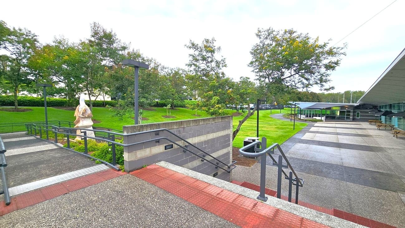 A paved outdoor area with a series of ramps and stairs in the foreground, featuring metal railings. On the left, a stone sculpture stands amidst green grass and trees. The background has a modern building with a sloped roof and glass windows. Several picnic tables are placed along the right side, near the building. The area is well-maintained, with neatly trimmed grass and scattered trees.