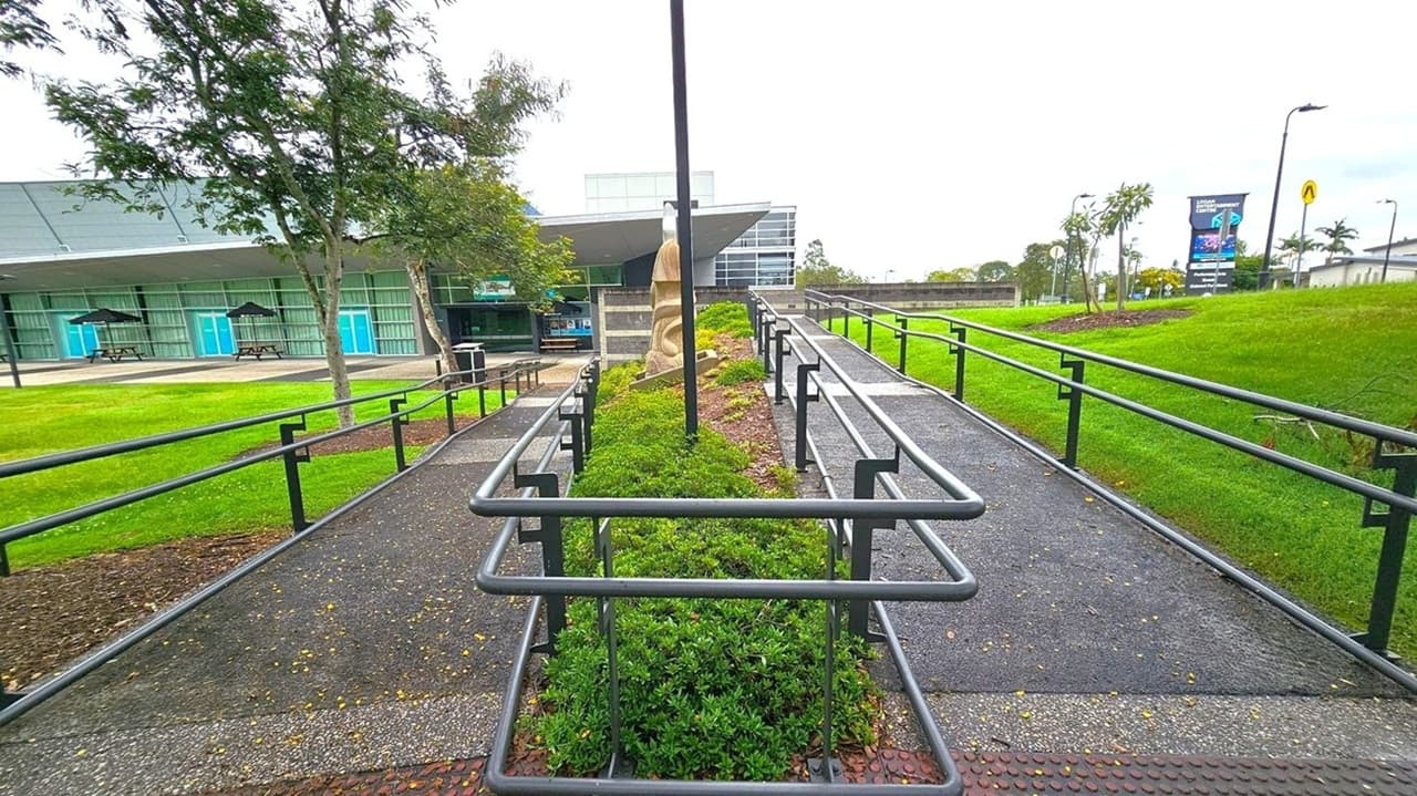 A pathway with metal railings runs through the centre, flanked by green shrubs. On the left, a building with large windows and blue doors is visible, with picnic tables and umbrellas outside. On the right, a grassy area with a few small trees and a sign in the distance. A sculpture is positioned in the middle of the pathway. The sky is overcast, and the ground is paved with small stones.