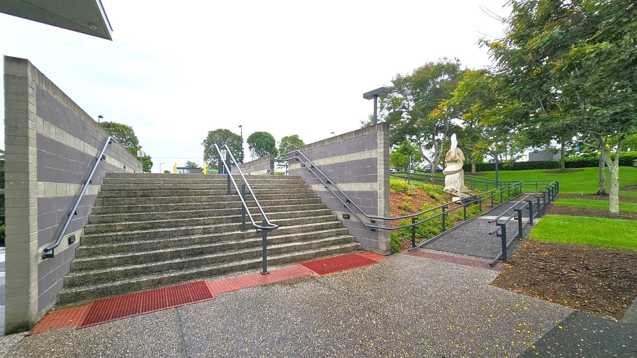 Wide stone steps with metal railings in the centre, flanked by grey brick walls. To the right, a sloped path with railings leads up, bordered by grass and trees. A statue stands on the right side of the path amidst greenery. The ground is speckled with small leaves or debris. In the background, more trees and a few lamp posts are visible.