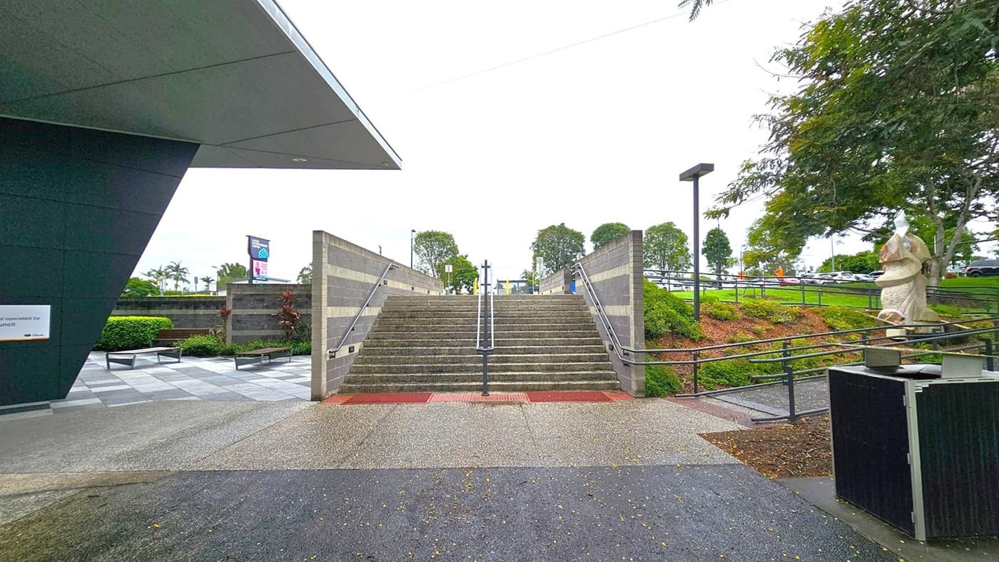 A set of wide stone steps with metal railings in the centre leads upwards, flanked by grey brick walls. On the left, a modern building with a dark overhanging roof and a sign is visible. To the right, a large sculpture stands near a tree, with a grassy area and a path leading to a car park in the background. The ground is a mix of concrete and grass, and there are benches and small shrubs on the left side.