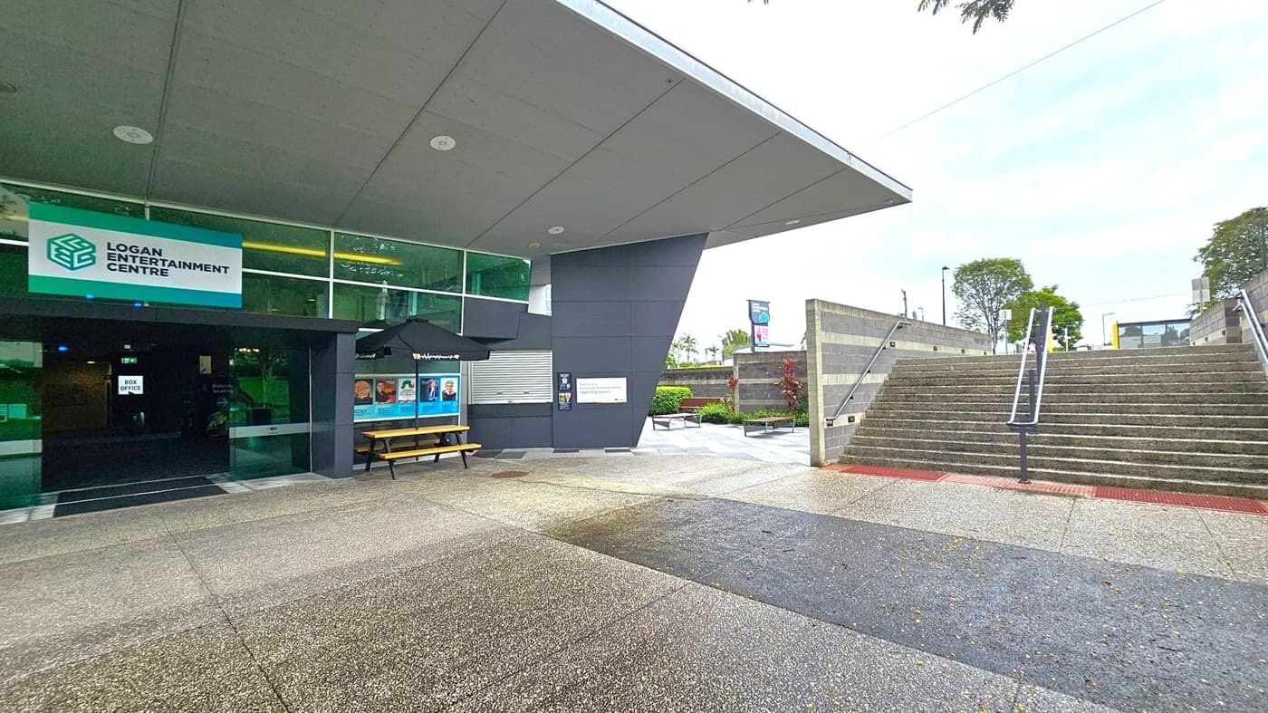 Entrance to a modern building with a sign reading "Logan Entertainment Centre" on the left. The entrance features glass doors and a box office. A bench and posters are under a black umbrella. On the right, a wide set of concrete stairs with metal railings leads upwards, flanked by grey walls. The ground is a mix of textured concrete and green patches. Trees and shrubs are visible in the background. The ceiling is flat and grey.