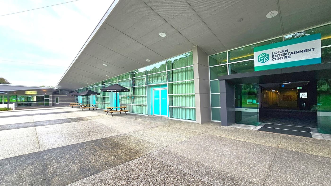 A modern building with a grey, angular roof and large glass windows. On the right, a sign reads "Logan Entertainment Centre" above a dark entrance with a "Box Office" sign inside. In the centre, blue double doors are flanked by glass panels with curtains. On the left, outdoor seating with wooden picnic tables and black umbrellas. The ground is paved with grey and speckled tiles.