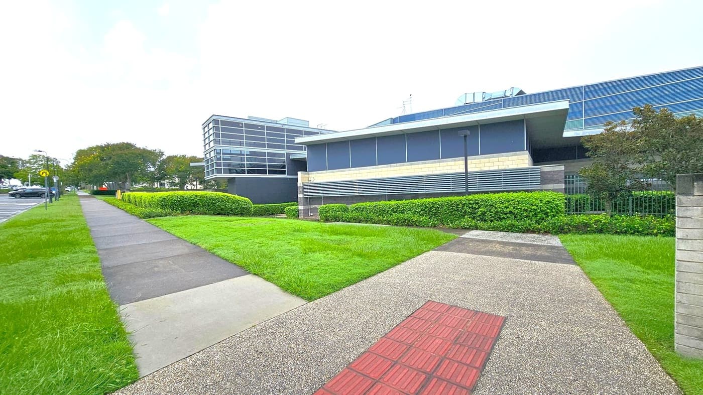 A modern building with a sleek design is on the right, featuring large windows and horizontal metal slats. In front, there's a well-maintained lawn bordered by neatly trimmed hedges. A paved path with a textured red section runs alongside the building. On the left, a concrete footpath is bordered by grass, leading towards a parking area with several cars and trees in the background. The scene is urban and orderly.