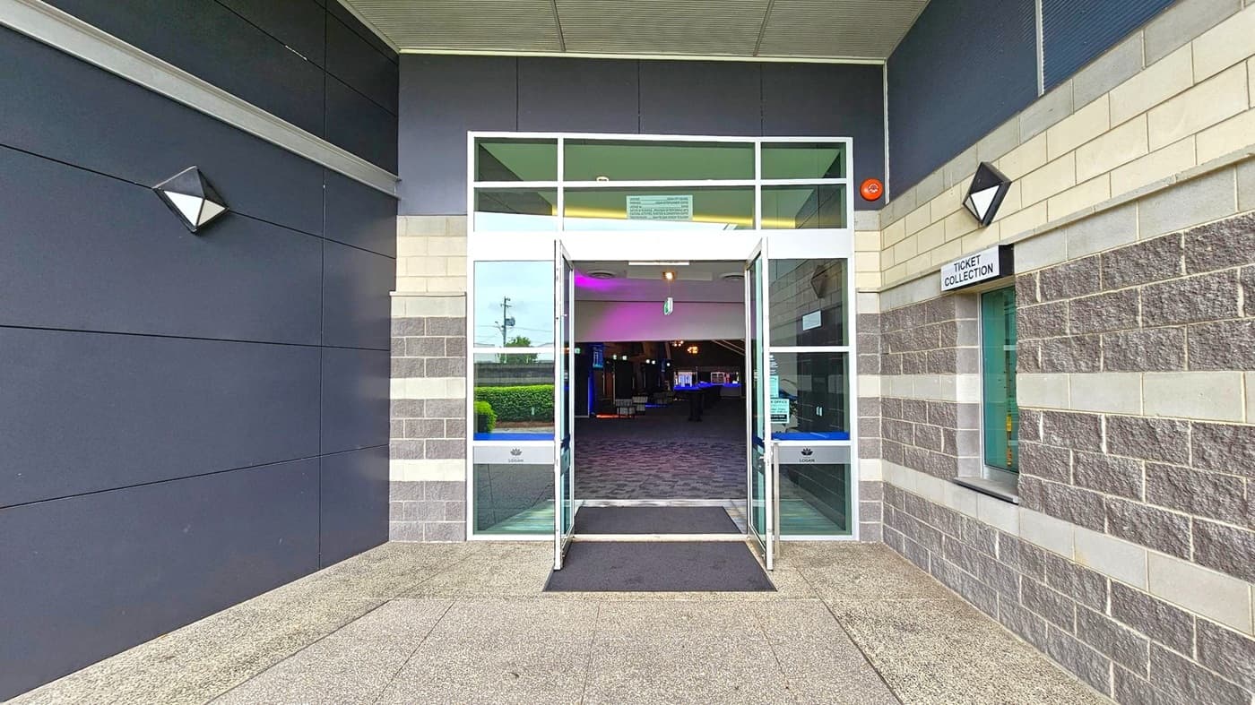 Entrance to a building with double glass doors open, leading to a dimly lit interior. The exterior walls are a mix of dark panels on the left and grey brick on the right. A sign above a window on the right reads "Ticket Collection". The floor is a speckled concrete surface, and there is a black mat in front of the doors. The ceiling is light-coloured with a grid pattern. Triangular wall lights are mounted on either side of the entrance.