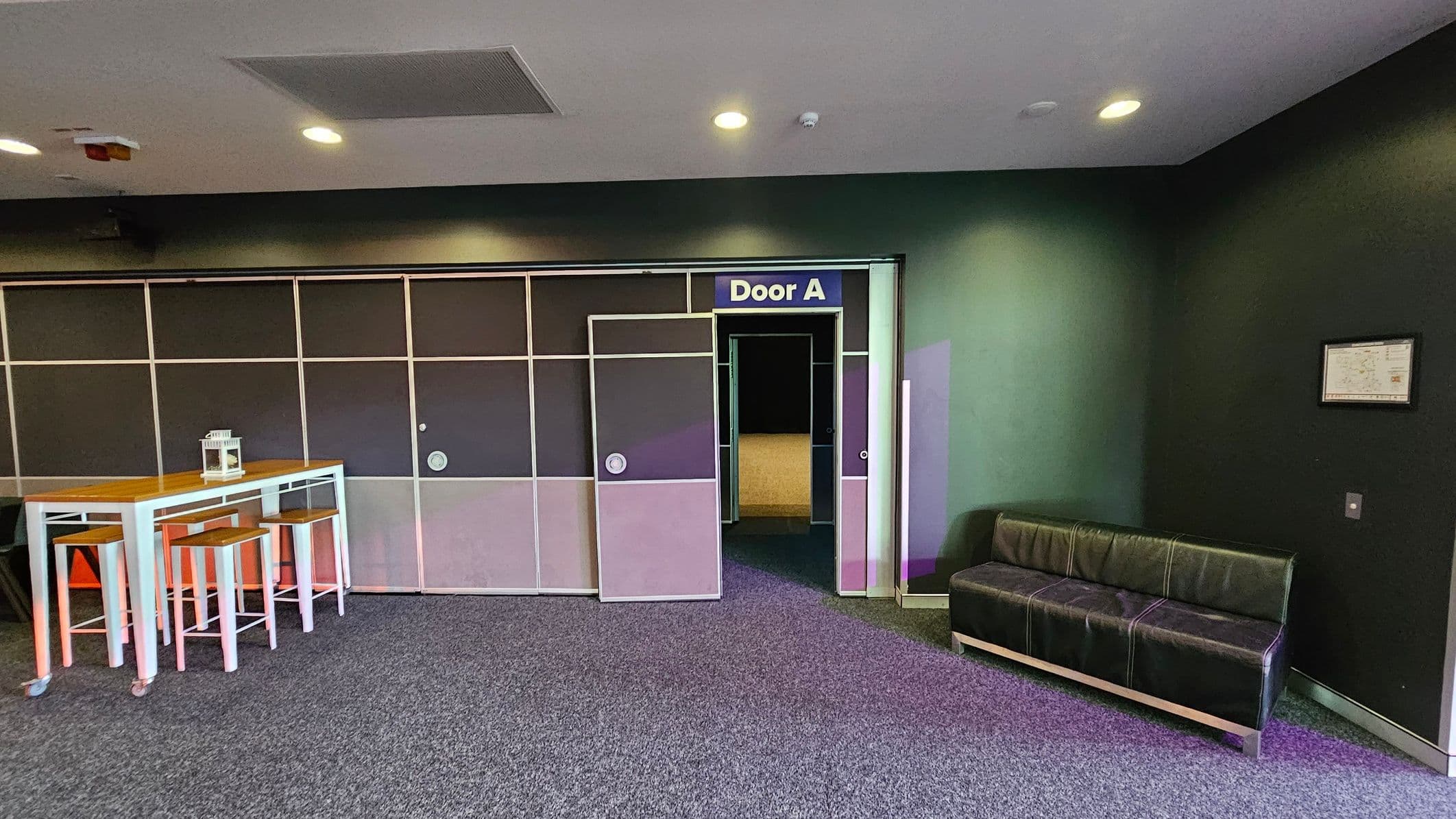 On the left, there is a tall wooden table with white metal legs, accompanied by three matching stools. A small lantern sits on the table. In the centre, a door labelled "Door A" is slightly open, revealing a dimly lit interior. On the right, a black leather sofa is positioned against a dark wall, with a framed item hanging above it. The floor is carpeted in grey, and the ceiling is fitted with recessed lights and a ventilation panel.