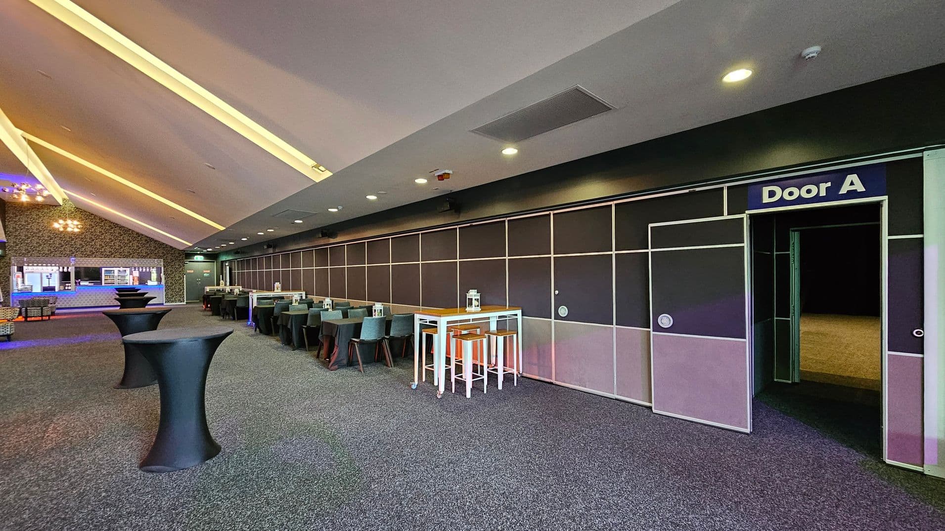 A spacious room with a grey carpeted floor and a ceiling featuring recessed lighting. On the right, a wall with a door labelled "Door A" leads to another area. In the centre, there are high tables with black covers and a white table with stools. On the left, a bar area with a counter and shelves is visible. The room has a modern design with a mix of dark and light tones.