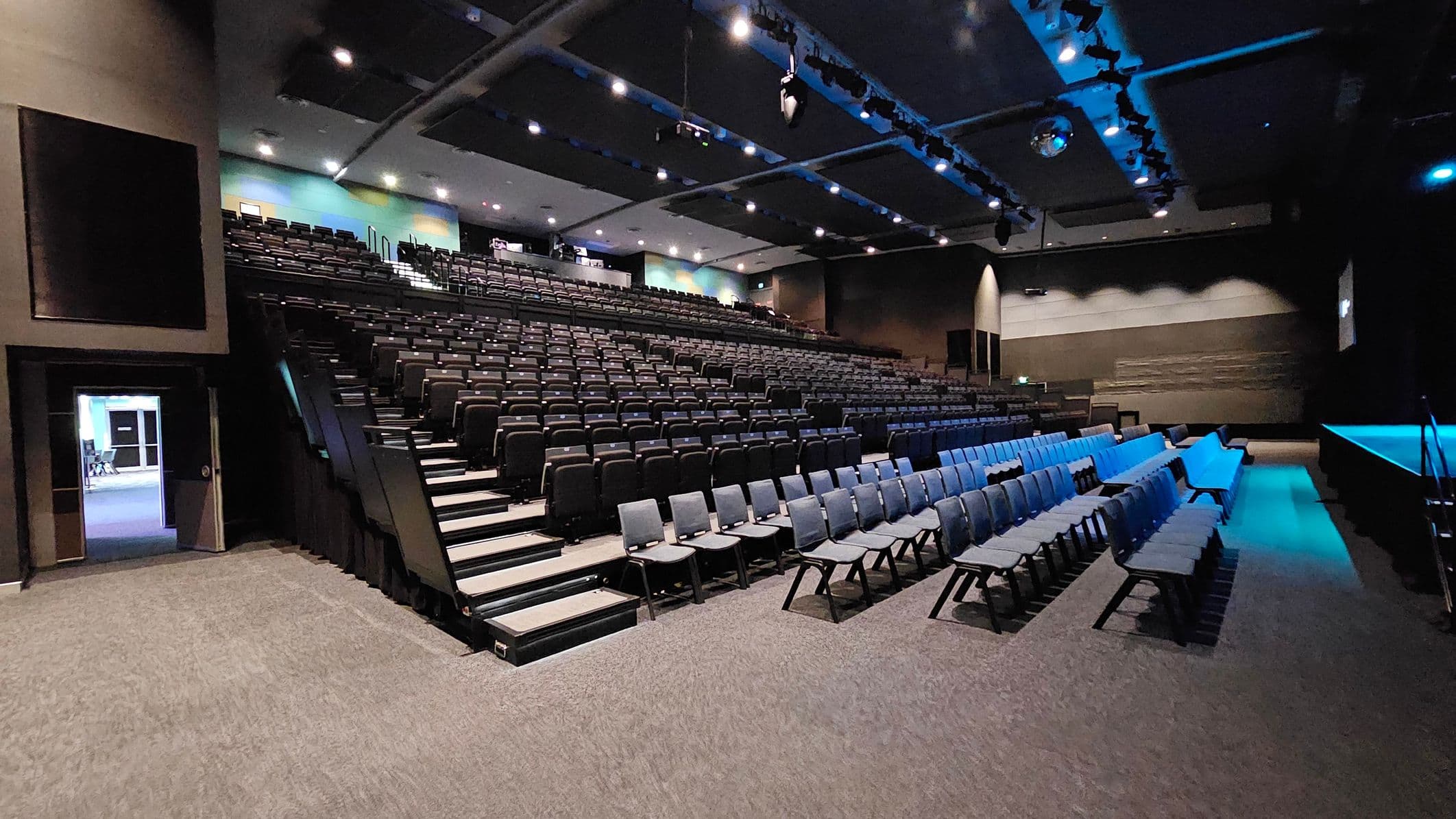A large auditorium with tiered seating on the left, featuring rows of black chairs. On the right, there are additional rows of chairs on a flat surface. The ceiling is equipped with spotlights and a disco ball, casting a blue hue over the room. The floor is carpeted in grey. A doorway on the far left leads to another room, and the walls are painted in dark tones with some light accents.