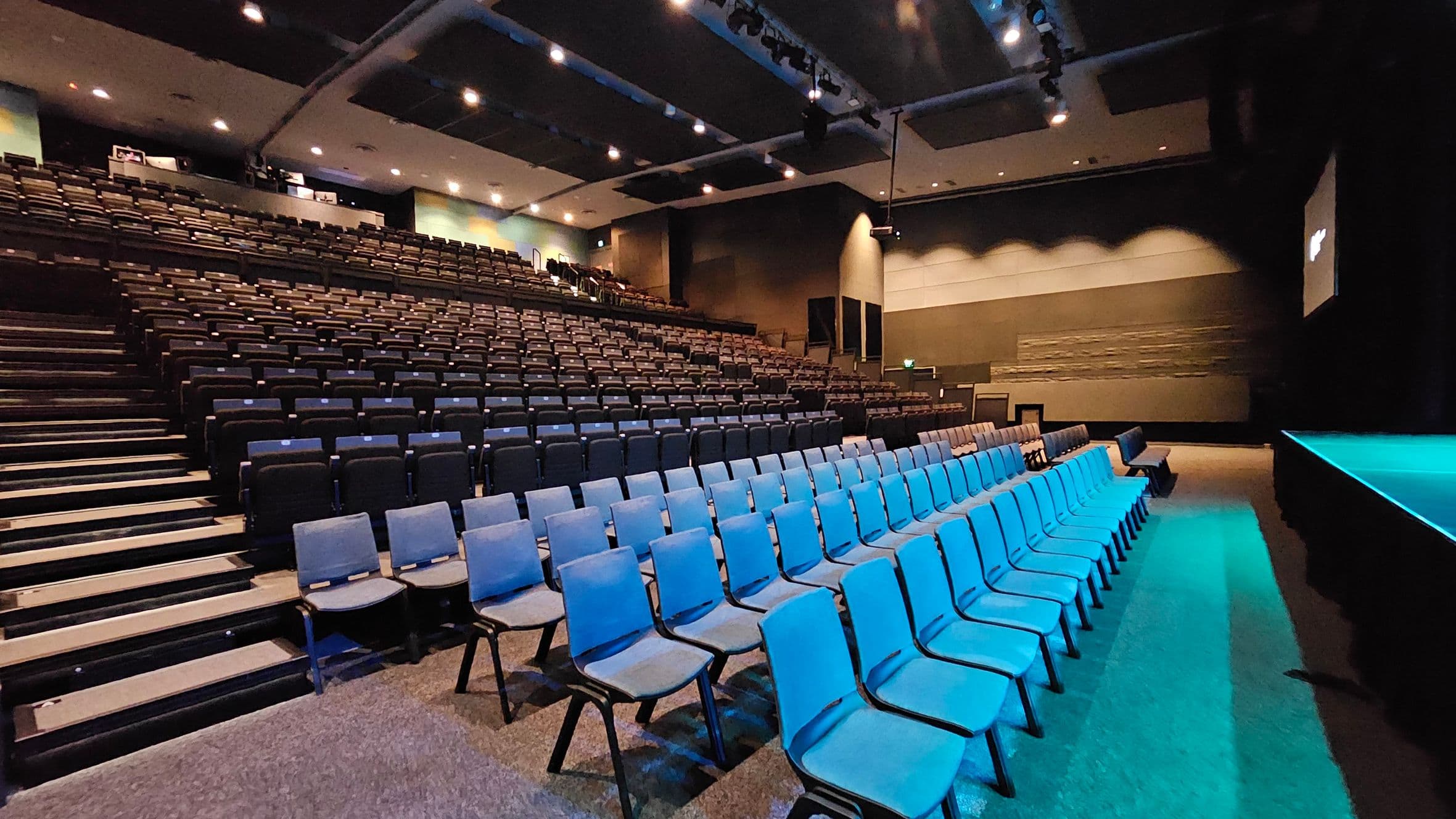 A large auditorium with tiered seating. On the left, rows of dark-coloured seats ascend towards the back. On the right, there are several rows of blue chairs on a flat surface. The stage is visible on the far right, illuminated with blue lighting. The ceiling is equipped with spotlights and acoustic panels. The walls are a mix of dark and light tones, with some textured sections. The floor is carpeted in a neutral colour.