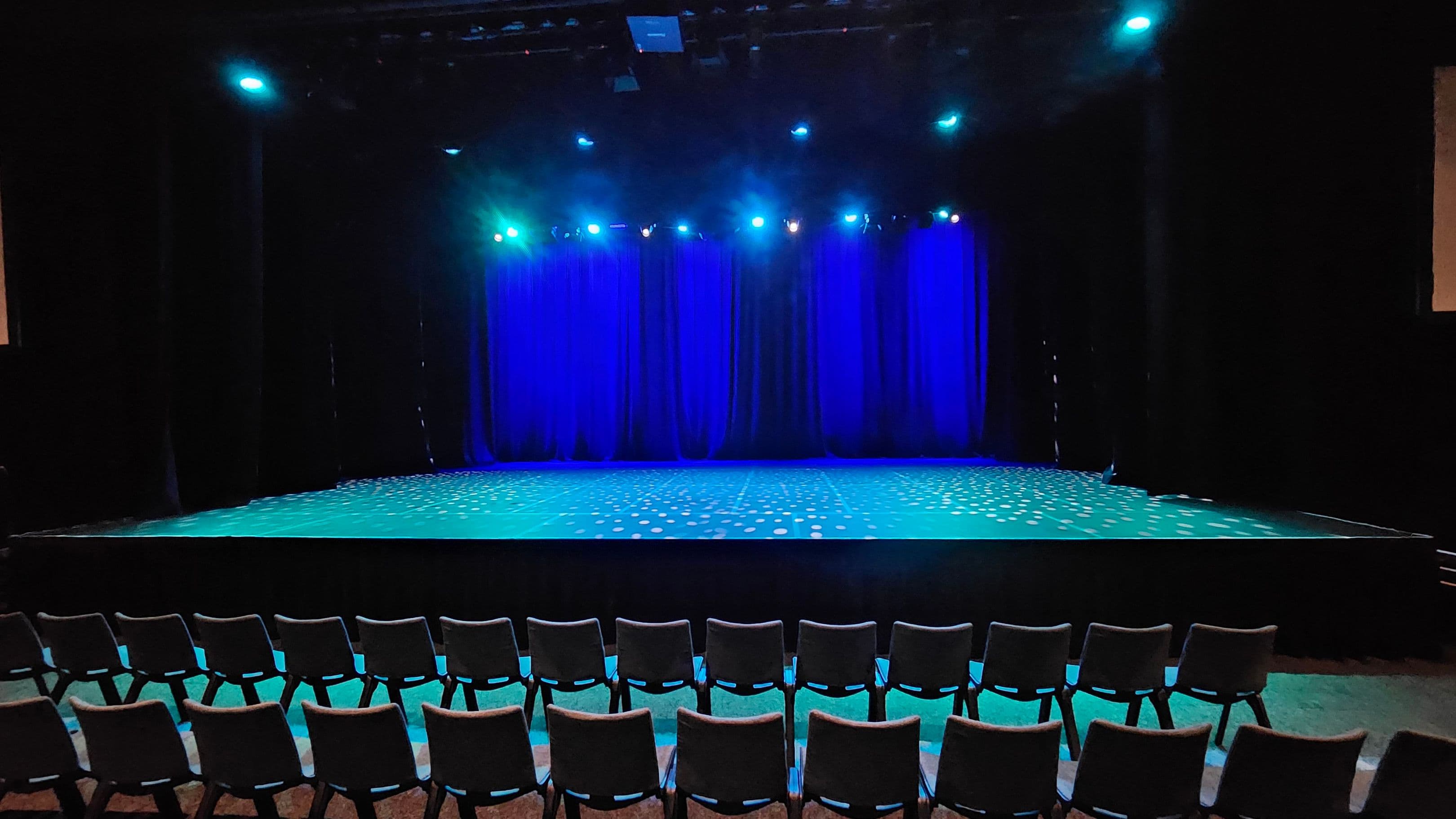 A theatre stage with a row of empty chairs in the foreground. The stage is illuminated with blue lighting, and the floor has a pattern of small white dots. Blue curtains hang in the background, and several spotlights are directed towards the stage. The ceiling is dark, with visible lighting fixtures. The overall atmosphere is dimly lit, with a focus on the stage area.