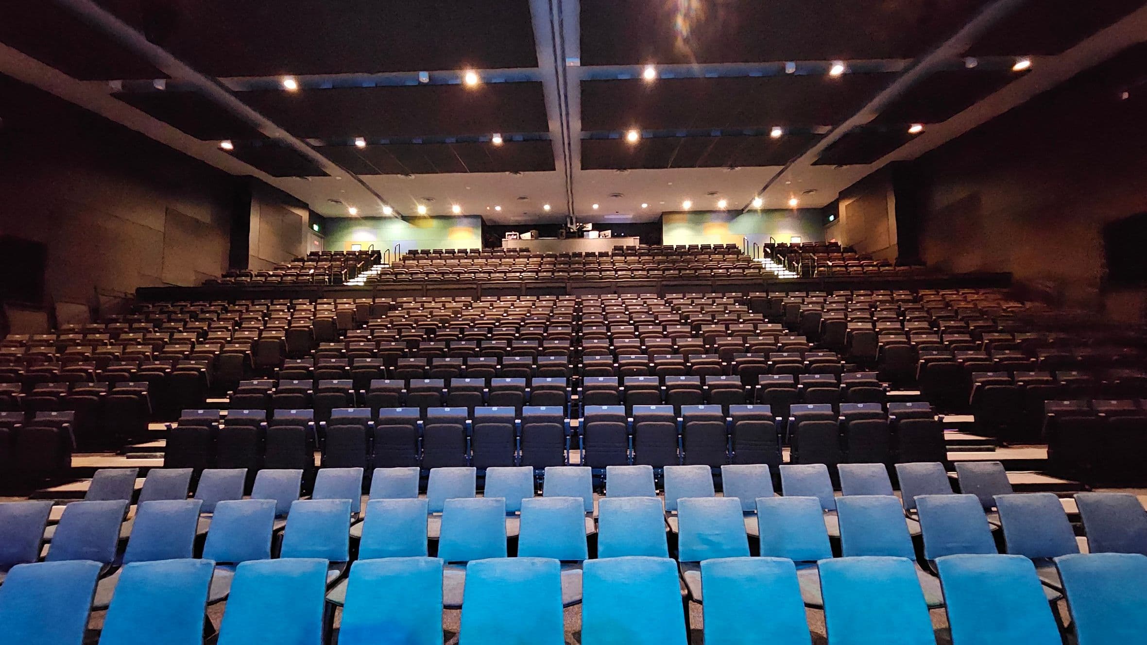 A large auditorium with rows of empty seats, predominantly in dark blue, facing the front. The seating is arranged in a tiered fashion, ascending towards the back. The ceiling is dark with evenly spaced recessed lighting. The walls are dark, and there are exits visible on either side towards the back. The floor is carpeted, and the overall atmosphere is spacious and organised.