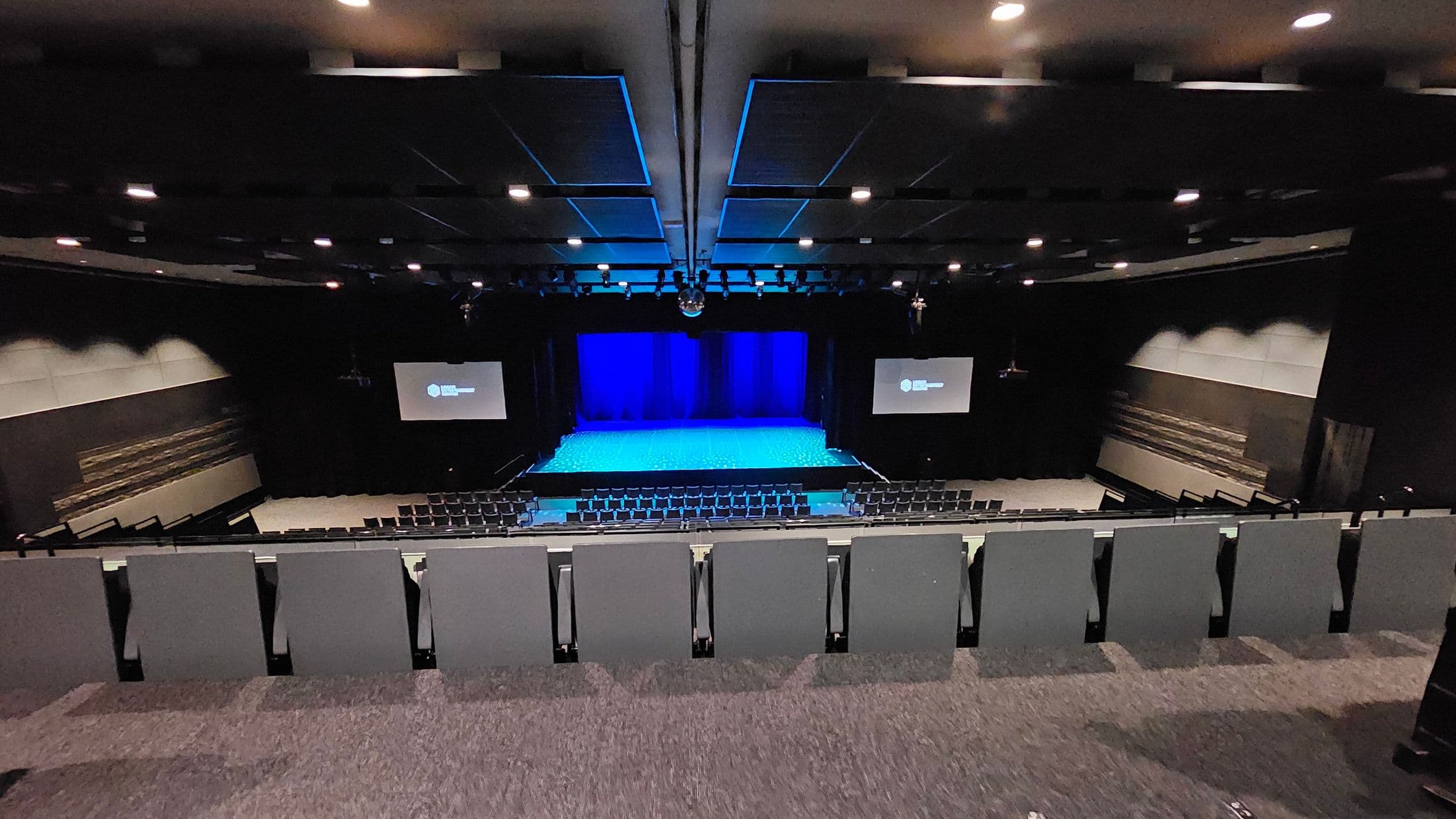 A large auditorium with tiered seating in the foreground, covered in dark fabric. The stage is in the centre, illuminated with blue lighting, and flanked by two large screens displaying text. The walls are lined with acoustic panels, and the ceiling features recessed lighting and sound equipment. The floor is carpeted in a dark colour, complementing the overall dimly lit atmosphere.