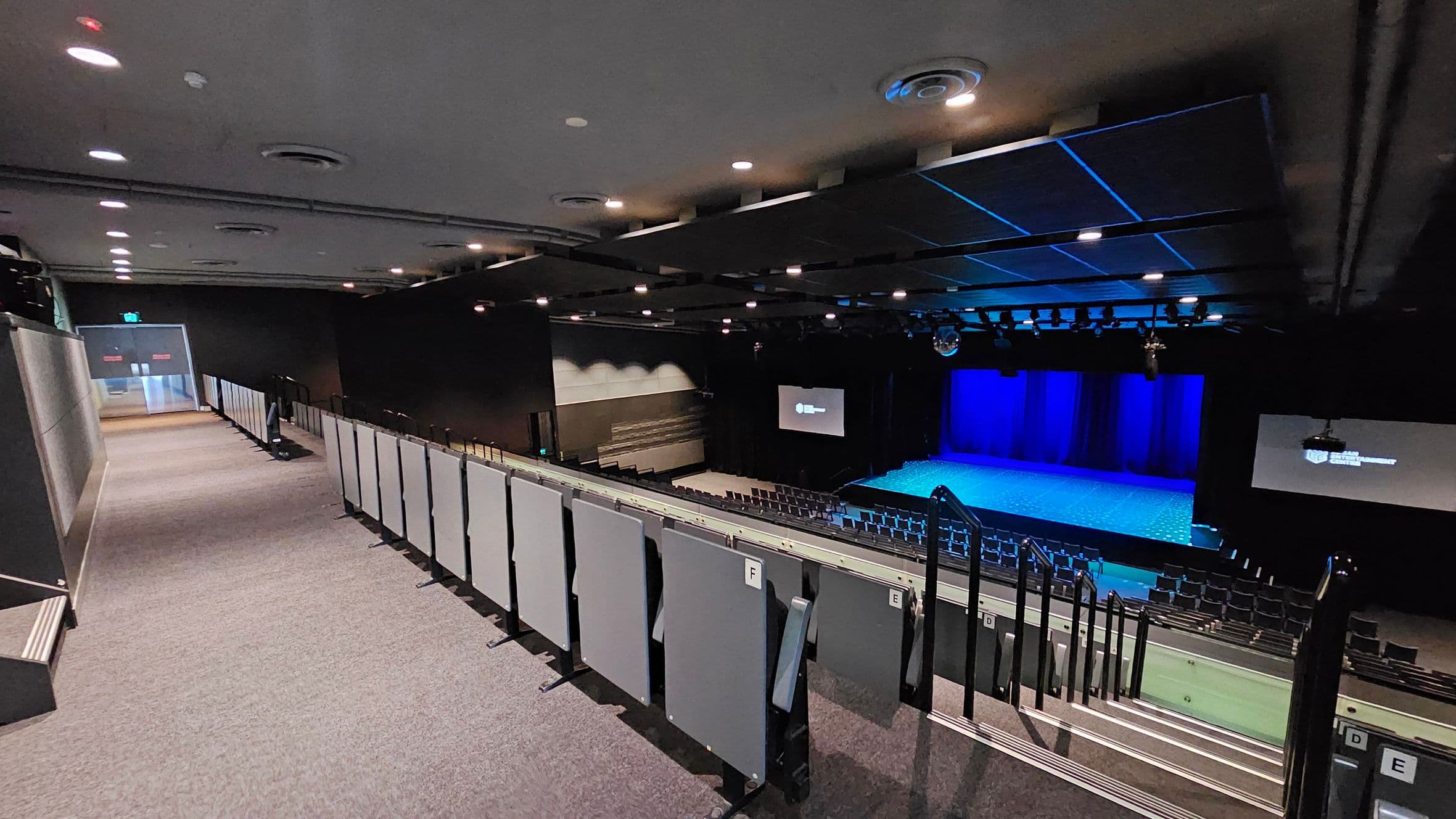 A theatre interior with a view from the upper seating area. On the left, a carpeted walkway leads to an exit door. The right side features tiered seating with grey panels and handrails. In the middle, a stage with a blue-lit backdrop is visible. Two screens flank the stage, displaying text. The ceiling is equipped with recessed lighting and ventilation ducts. The floor is carpeted, and the overall atmosphere is dimly lit.