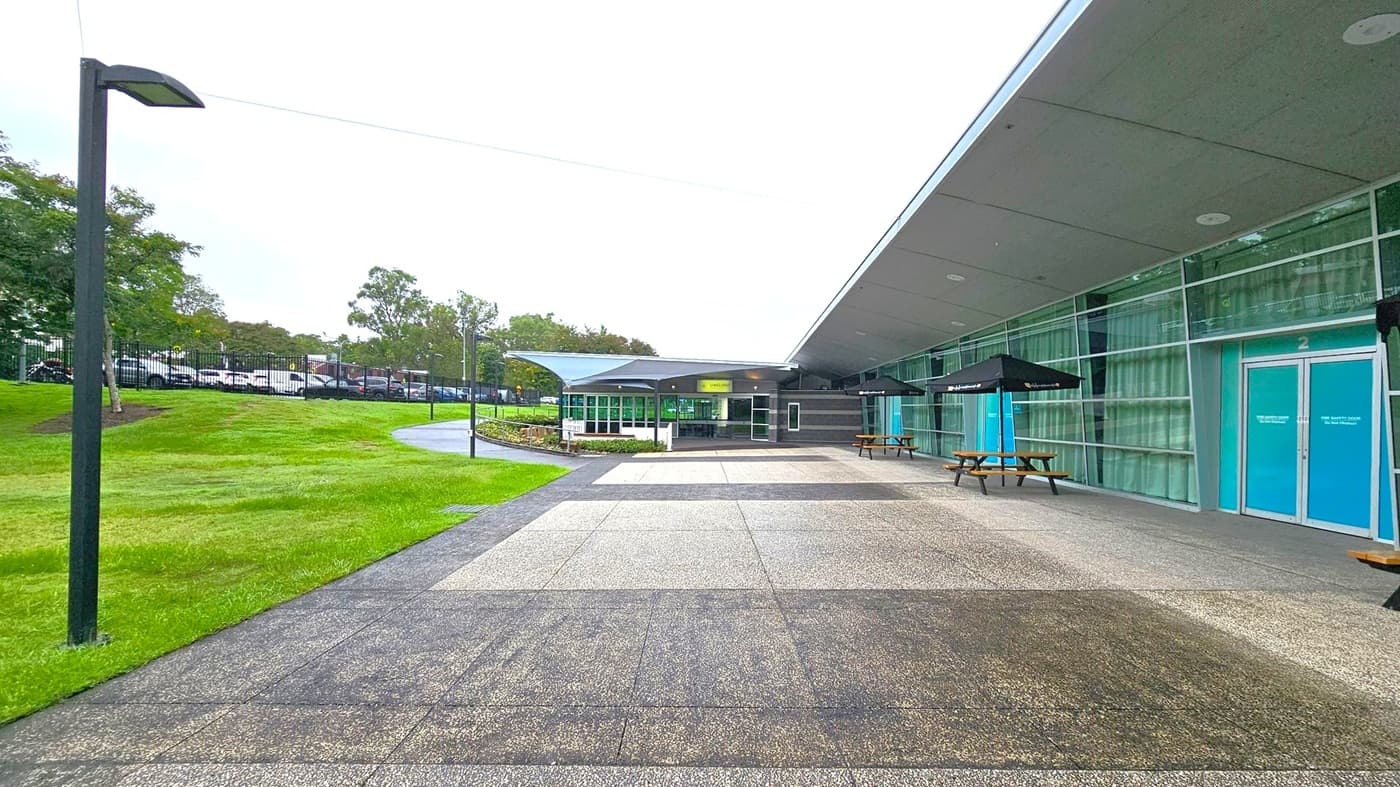 A modern building with a glass facade on the right, featuring blue doors and large windows. Two picnic tables with black umbrellas are placed along the building. The foreground has a paved area leading to a grassy lawn on the left, with a path and several trees. A row of parked cars is visible in the background, separated by a fence. The ceiling of the building extends outward, providing some shelter over the entrance area.