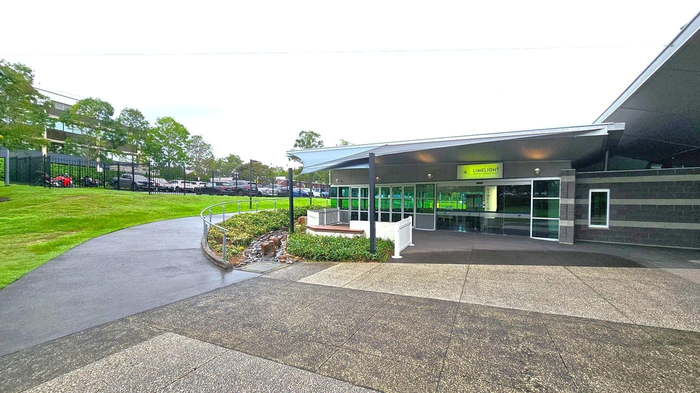 A modern building with a sign reading "LIMELIGHT" above the entrance, featuring large glass doors and windows. The structure has a sloped roof and is surrounded by a paved area with a mix of grey and green tiles. To the left, a curved path leads up a grassy hill, bordered by a metal railing and small shrubs. In the background, there are trees and a fenced parking area with several cars.