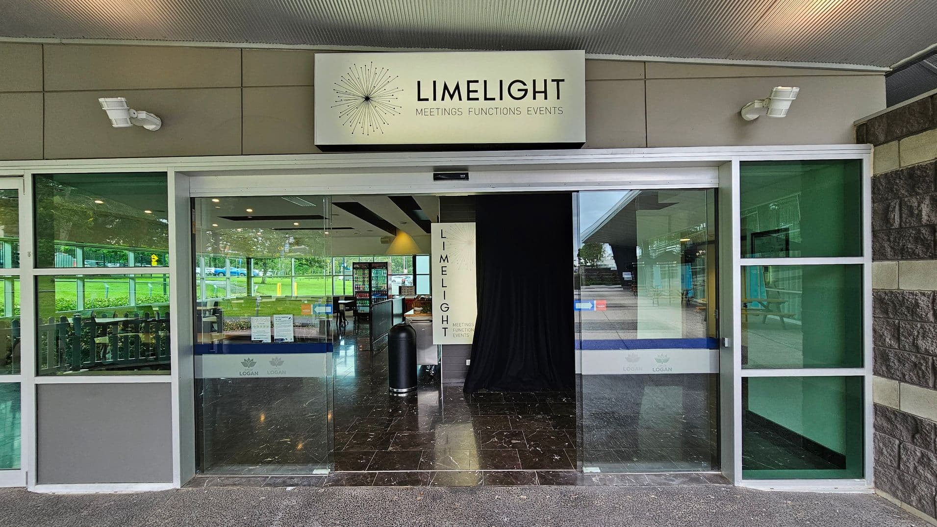 A glass entrance with a sign above reading "LIMELIGHT MEETINGS FUNCTIONS EVENTS". The entrance has large glass doors and windows with green-tinted panels on either side. Inside, there is a black curtain and a glimpse of a seating area with tables and chairs. The floor is dark and glossy, reflecting the interior lights. The exterior has a textured stone wall on the right and a grey ceiling with lights.