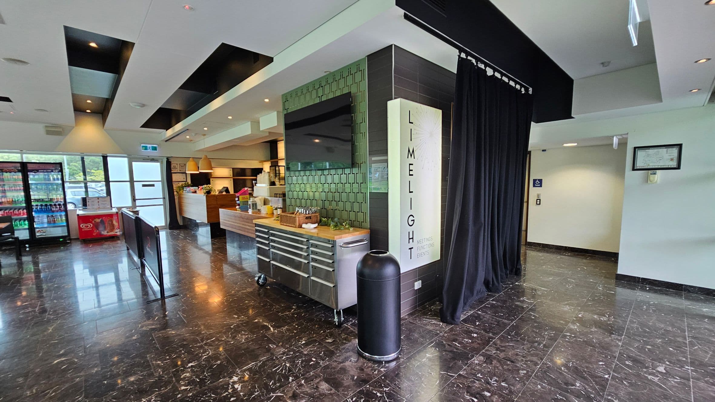 A modern interior with a glossy black marble floor. On the left, there are large windows and a fridge stocked with drinks. In the centre, a counter with wooden accents and hanging lights is visible. A black curtain and a sign reading "LIMELIGHT" are on the right, next to a black cylindrical bin. The ceiling features recessed lighting and black panels.