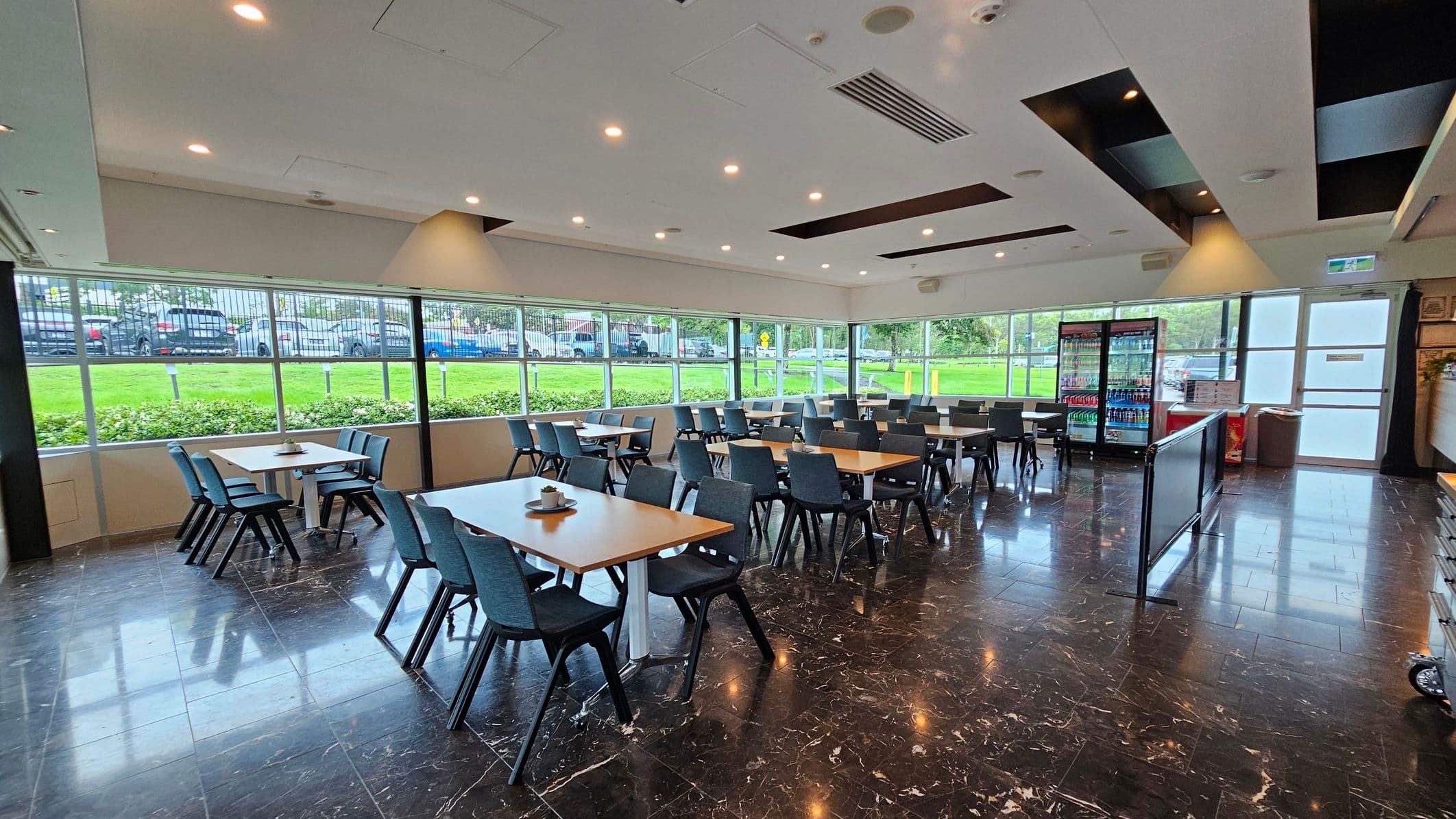 A spacious dining area with several wooden tables and black chairs arranged neatly. Large windows on the left and background allow natural light to illuminate the room, offering a view of a grassy area and parked cars outside. On the right, there are refrigerated display units stocked with beverages. The floor is dark with a polished finish, and the ceiling features recessed lighting and air vents.
