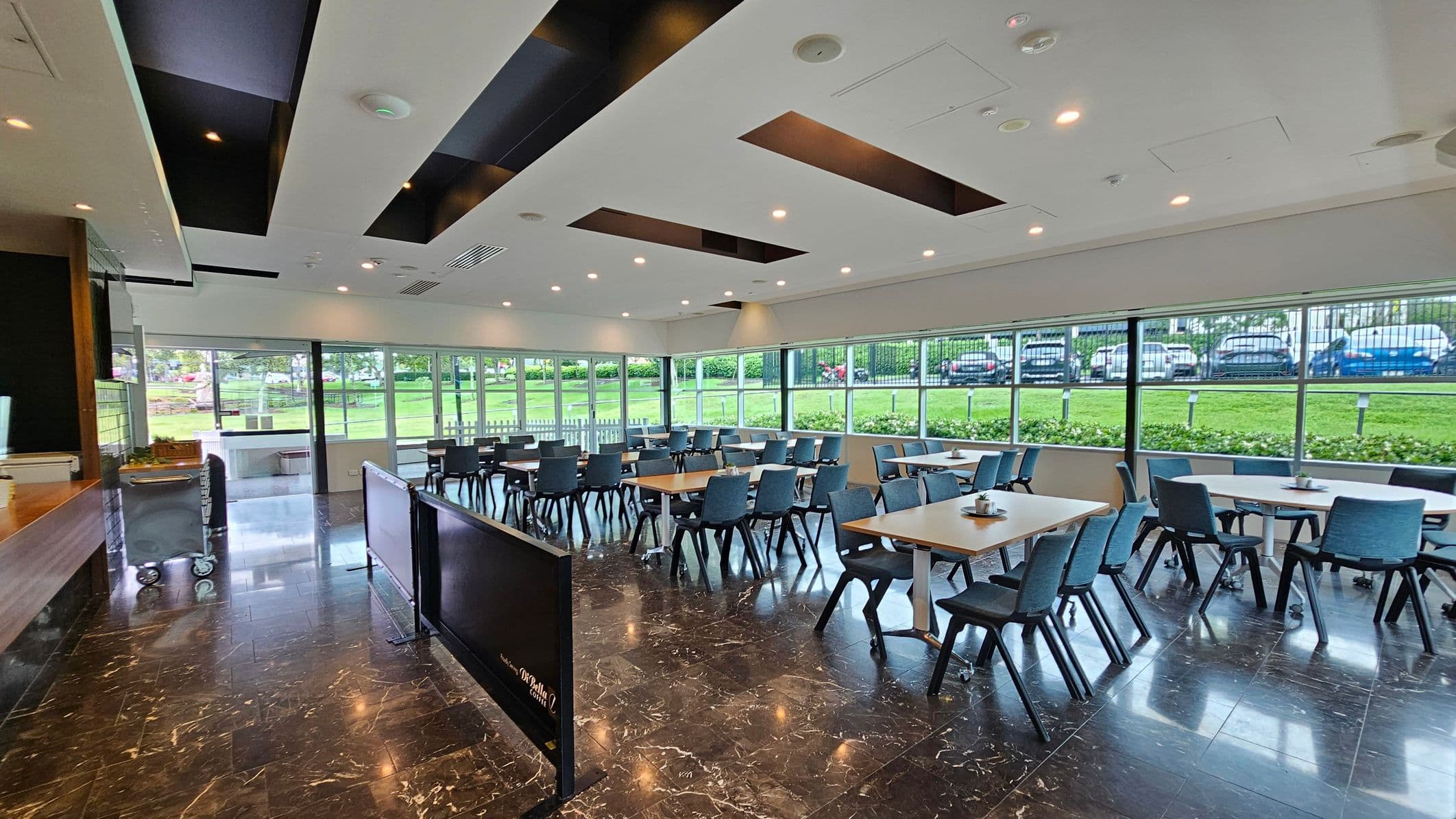 A spacious dining area with dark marble flooring and a modern ceiling featuring recessed lighting and black accents. On the left, there's a counter with a trolley nearby. The right side has several tables and chairs arranged neatly. Large windows in the background offer a view of a grassy area and parked cars outside.