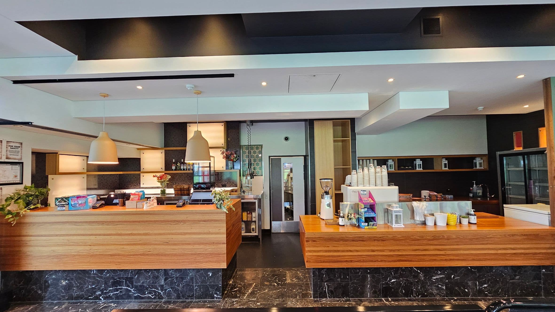 A modern café counter with a wooden finish. On the left, there are two large pendant lights above the counter, which displays snacks and a cash register. A plant and some flowers add a touch of greenery. In the centre, a door leads to the back area. On the right, a coffee machine and cups are visible, with shelves holding various items. The floor is dark marble, and the ceiling is white with recessed lighting.