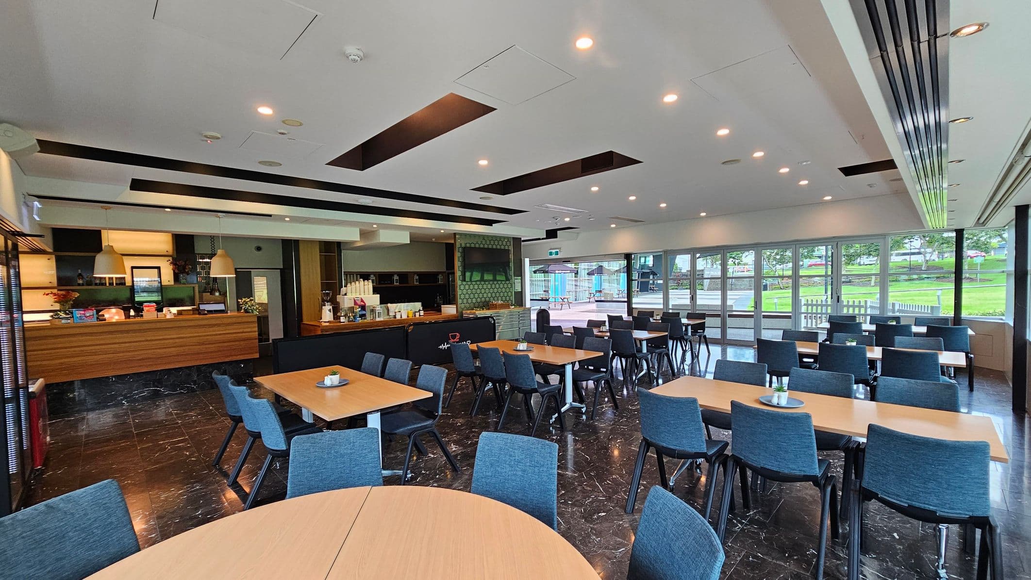 A spacious dining area with several wooden tables and blue chairs arranged neatly. On the left, there's a counter with pendant lights above, displaying various items and a coffee machine. The floor is dark marble, and the ceiling is white with recessed lighting. Large windows on the right offer a view of a green outdoor area with trees and picnic tables. The room is well-lit and modern in design.