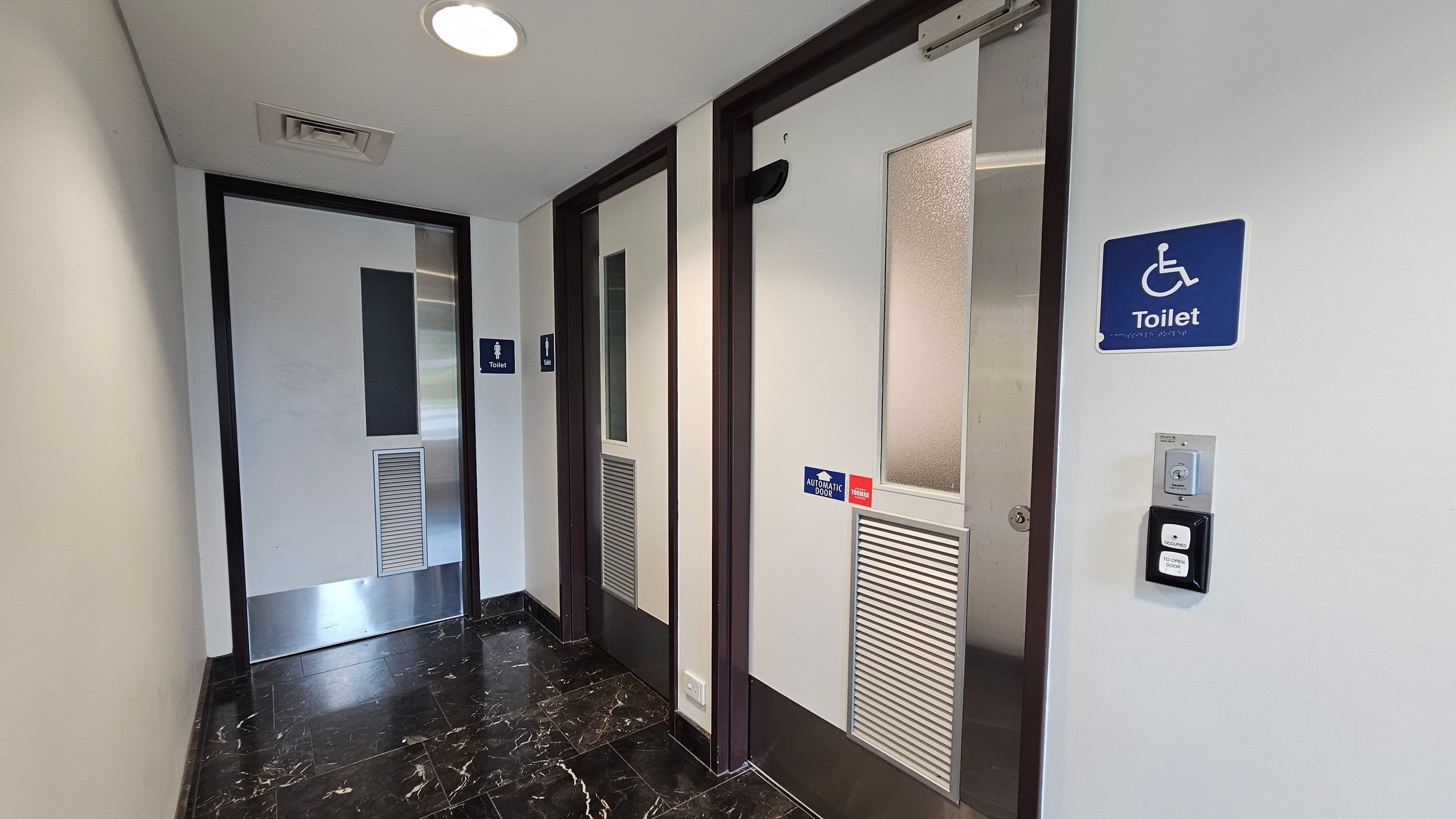 A corridor with two toilet doors on the right, one marked with a wheelchair symbol for accessibility. Both doors have frosted glass panels and ventilation grilles. The accessible toilet has an automatic door sign and a button for operation. The floor is black marble with white veining, and the ceiling is white with recessed lighting and a vent. On the left, a plain wall leads to another door at the end of the corridor.