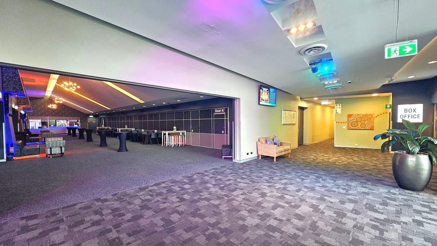 A spacious interior with a patterned carpeted floor. On the left, an open area with high tables and chairs, illuminated by modern chandeliers. A sign reads "Door A". On the right, a hallway with a box office sign, a potted plant, and a beige sofa with colourful cushions. The ceiling features recessed lighting and air vents. An exit sign is visible above the hallway.