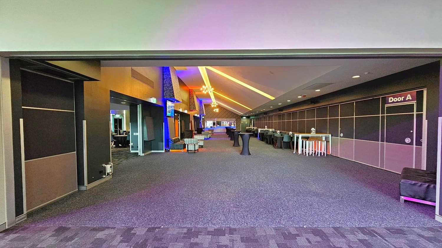 A spacious interior with a high, slanted ceiling featuring recessed lighting and chandeliers. The floor is carpeted in a dark grey pattern. On the left, there are open doorways and seating areas with armchairs. On the right, a long wall with a sign for "Door A" and several high tables with stools. In the background, a bar area is visible, and the space is illuminated with colourful lighting accents.