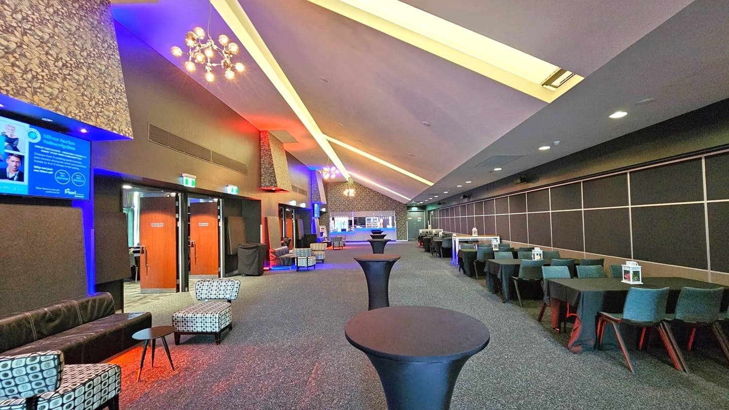 A spacious, modern lounge area with a high, slanted ceiling featuring recessed lighting. On the left, there are patterned chairs and a black leather sofa near wooden doors. A digital screen is mounted on the wall. The right side has a row of tables with teal chairs, each adorned with a small decorative item. The floor is carpeted, and several tall, round cocktail tables are positioned along the centre. The lighting creates a warm, inviting atmosphere with blue and red accents.