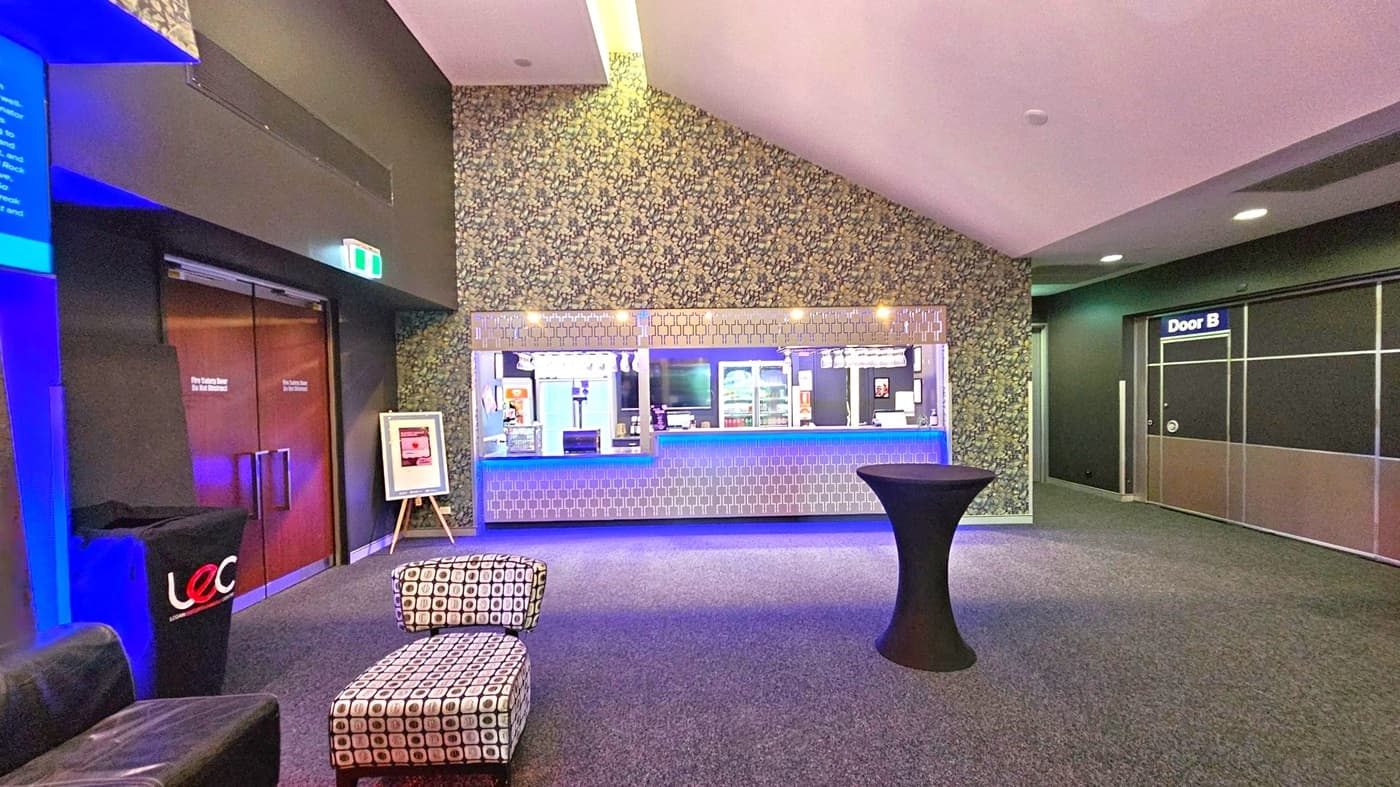 A modern interior with a patterned wall in the centre featuring a counter with a geometric design and blue lighting. Above the counter, glasses hang upside down. To the left, there are double wooden doors with a framed sign on an easel nearby. A patterned ottoman and a black cocktail table are in the foreground. On the right, a door labelled "Door B" is visible. The floor is carpeted, and the ceiling has recessed lighting.
