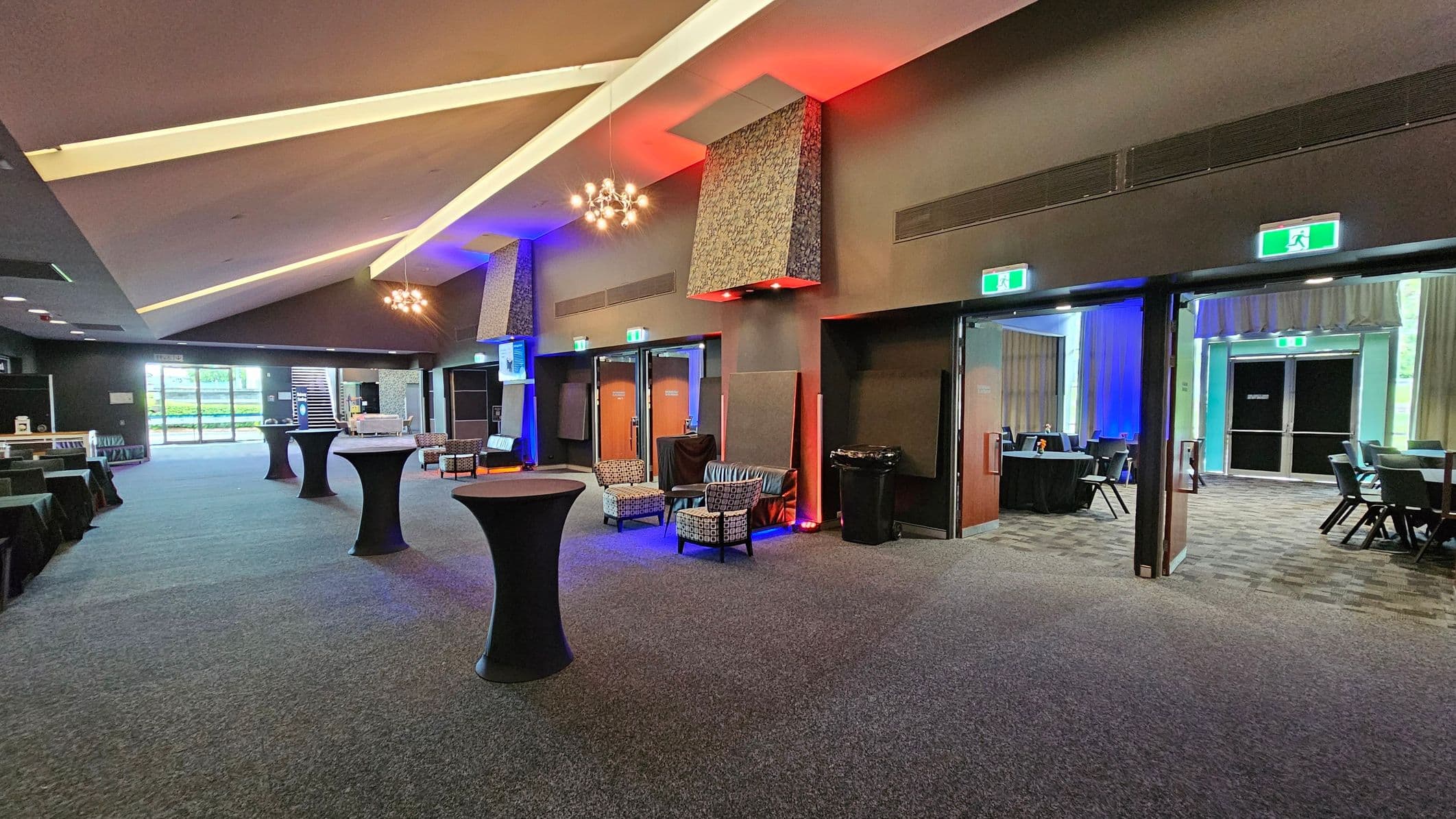 A spacious interior with a high, angled ceiling featuring modern light fixtures. The floor is carpeted in a grey tone. On the left, there are tables covered with black cloths and a large window with a view of greenery. In the centre, tall cocktail tables are scattered, and a seating area with patterned chairs is visible. On the right, there are open doors leading to another room with more tables and chairs. The walls are adorned with decorative panels and illuminated with coloured lights.
