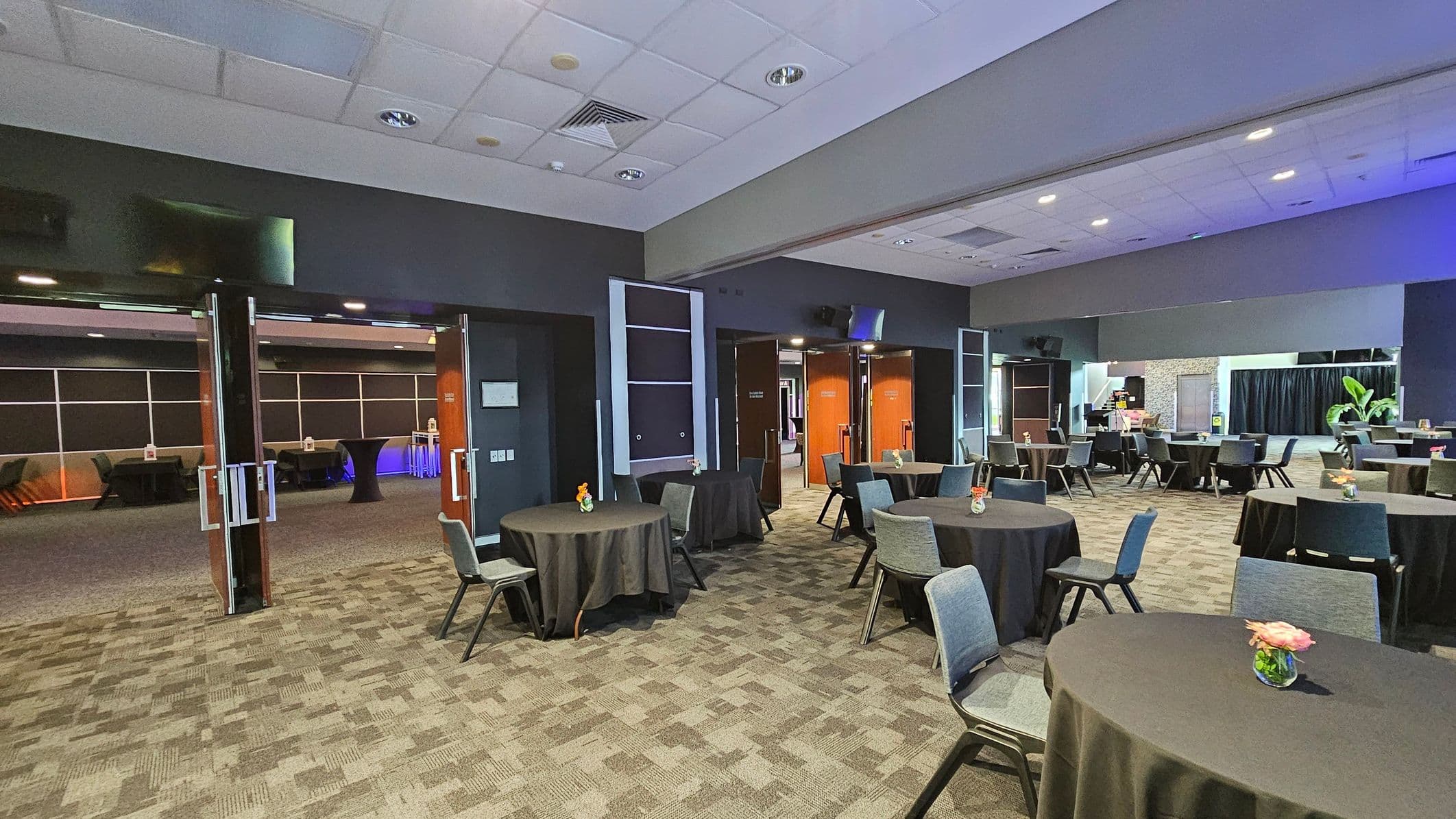 A spacious conference room with a patterned carpeted floor and a white panelled ceiling. Round tables covered with black tablecloths are scattered throughout, each adorned with a small floral centrepiece. Grey chairs are arranged around the tables. In the background, open double doors lead to another room with more tables and a blue-lit area. Large plants and a black curtain are visible in the far right corner.