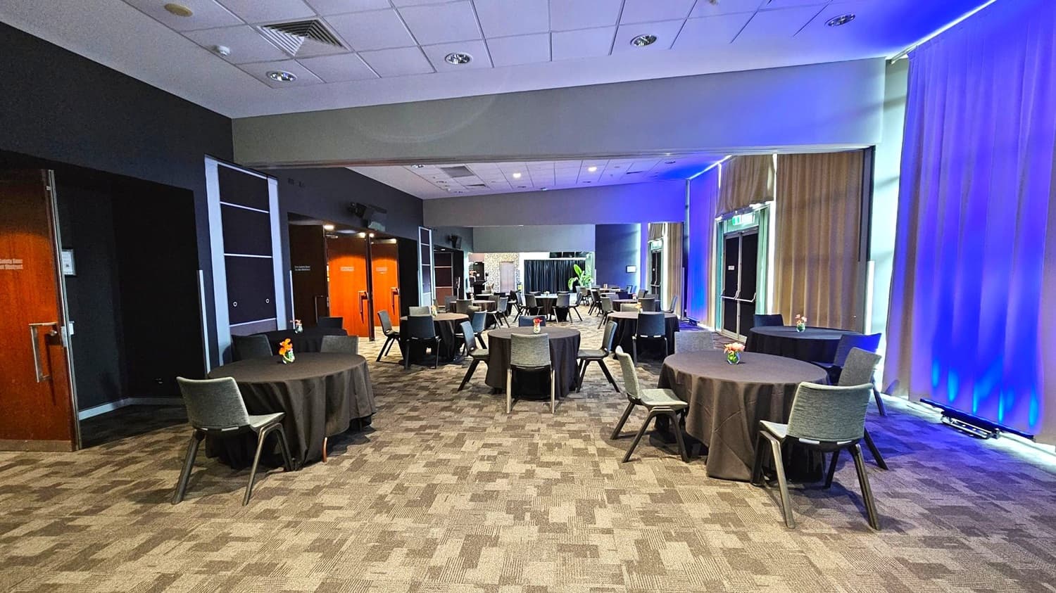 A conference room with round tables covered in black tablecloths, each with a small floral centrepiece. Grey chairs are arranged around the tables. The left side has wooden doors and dark walls, while the right side features large windows with beige curtains illuminated by blue lighting. The ceiling is white with recessed lighting, and the floor is carpeted in a patterned design. In the background, there is a plant and more seating.