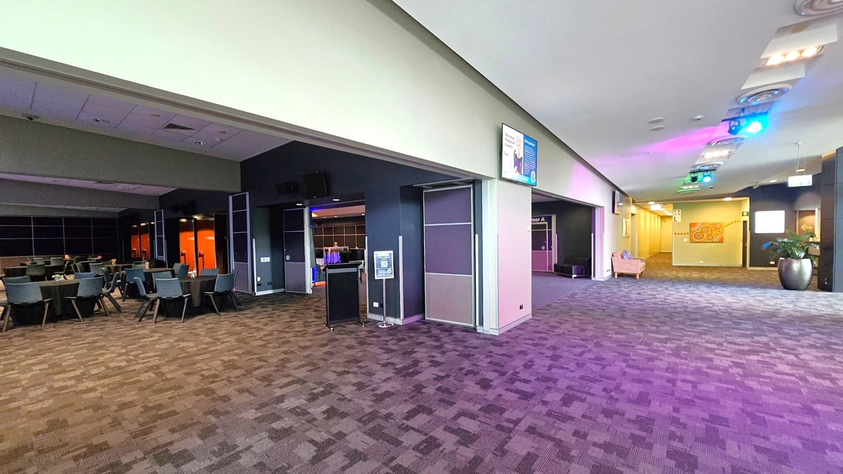 A spacious interior with a patterned carpet floor. On the left, there are tables and chairs arranged in a dining or conference style. The right side features a wide corridor with a potted plant and a beige armchair. The ceiling has recessed lighting and colourful spotlights. Walls are adorned with signs and a decorative mural. The area appears to be a multifunctional space, possibly in a conference centre or hotel.