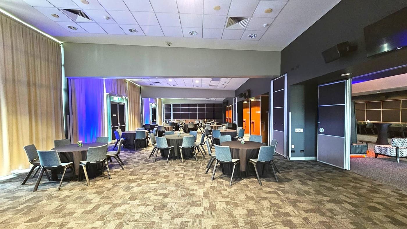 A spacious conference room with round tables covered in black tablecloths, each surrounded by grey chairs. The left side features beige curtains with blue lighting accents. The right side has dark walls with open sliding doors leading to another area with patterned chairs. The ceiling is white with recessed lighting and air vents. The floor is carpeted in a geometric pattern of brown and grey tones.