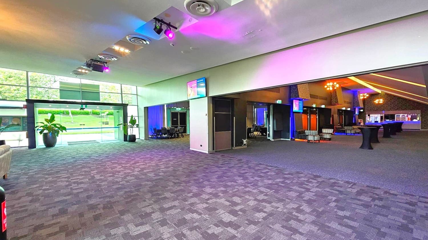A spacious interior with a patterned carpeted floor. On the left, large glass doors lead outside, flanked by potted plants. The ceiling features circular vents and colourful lights. On the right, an open area with tables and chairs, illuminated by warm lighting. A bar is visible in the background. Monitors are mounted on the walls, and a sofa is partially visible on the left.
