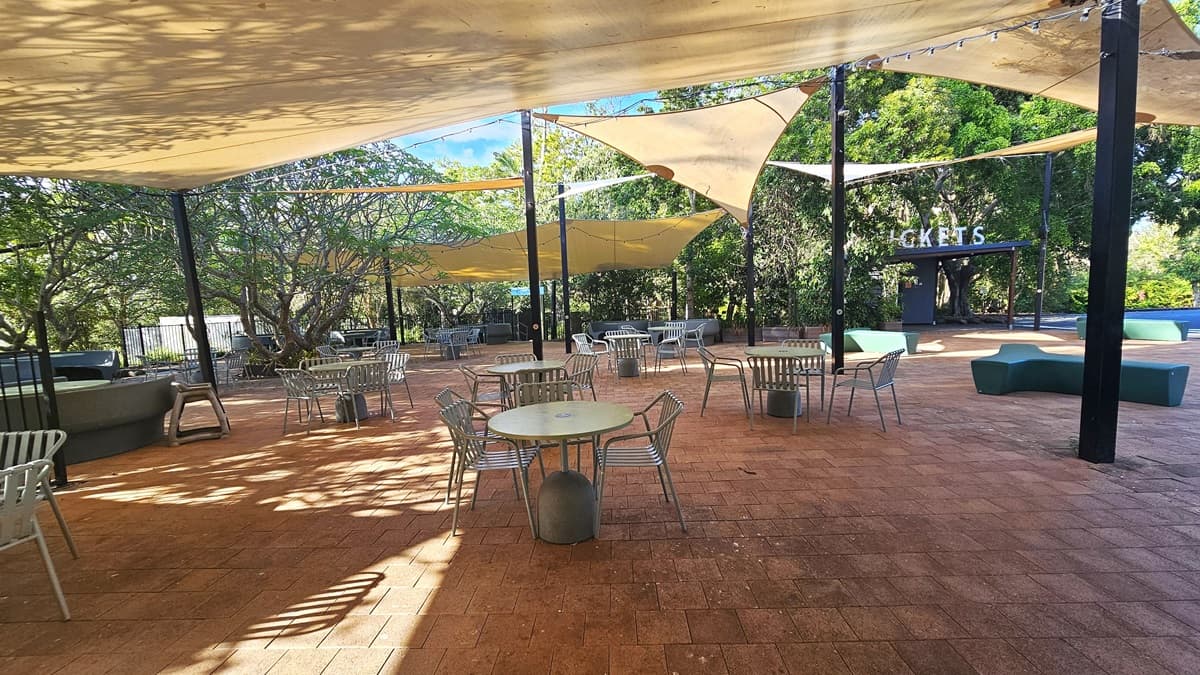 A closer look at the outdoor seating area under the numerous cream shades. Several chairs and tables are clearly organized. Mini light bulbs are prominently hanging adjacent to the cream shades. Benches with unique designs are scattered on the right side area. Further ahead on the right, the ticketing office signage is prominent.